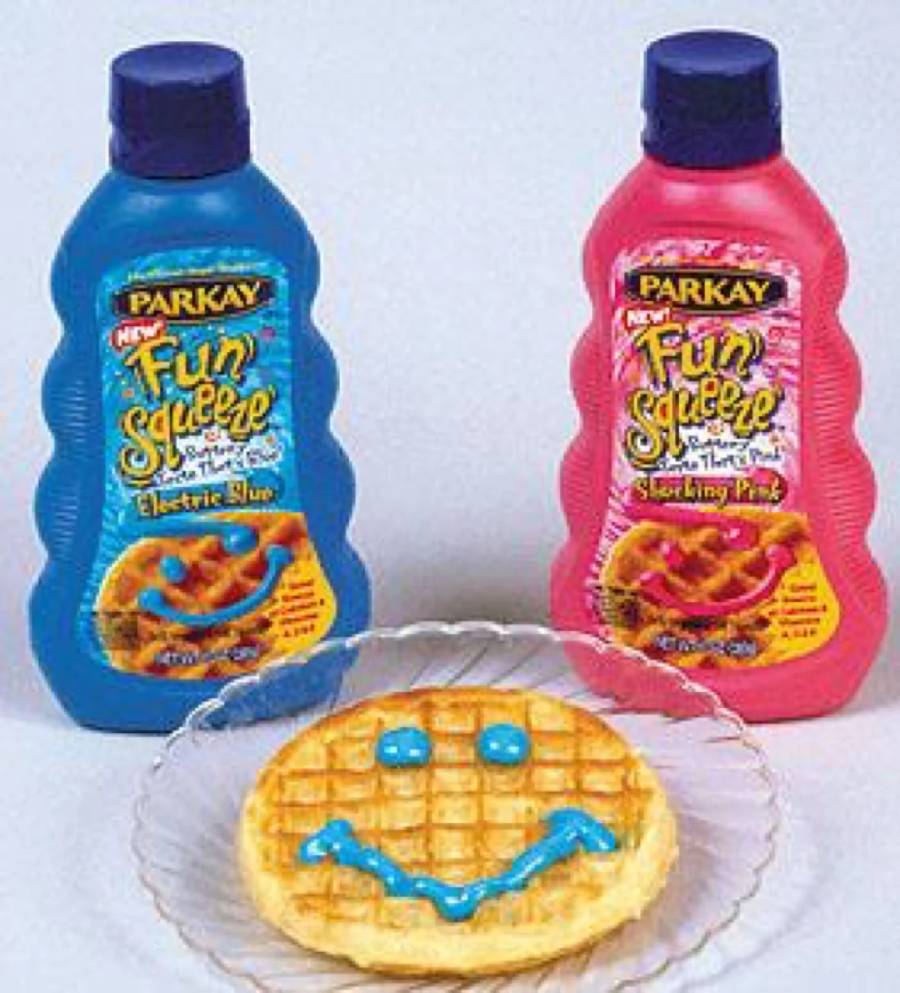A waffle on a clear plate is decorated with a blue smiley face, next to bottles of Parkay Fun Squeeze buttery spreads in blue (“Electric Blue”) and pink (“Shocking Pink”) varieties.