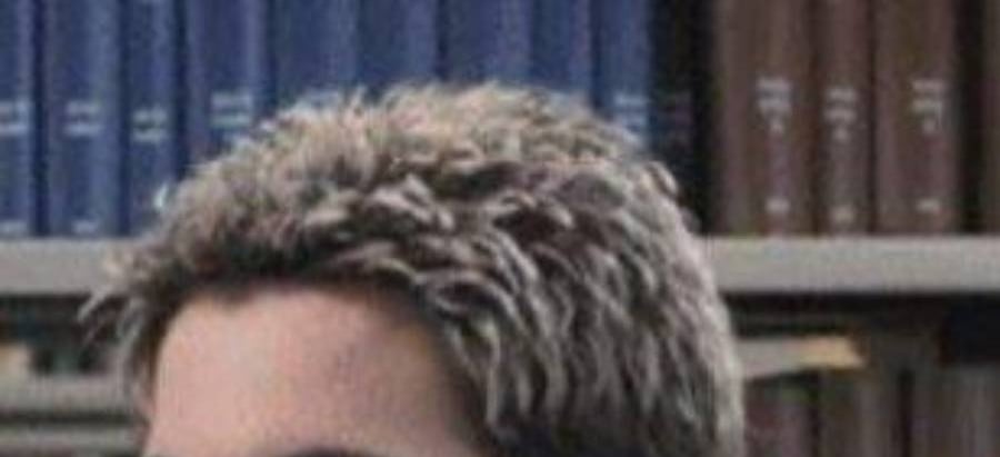 A person with short, spiked hair with light tips is in front of bookshelves filled with blue and brown books.