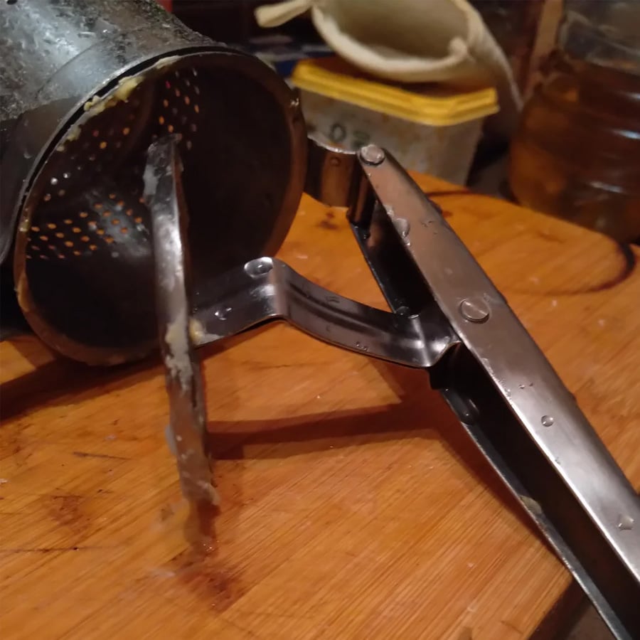 A close-up of a metal potato ricer on a wooden cutting board, with bits of food visible on its perforated plate and handle.