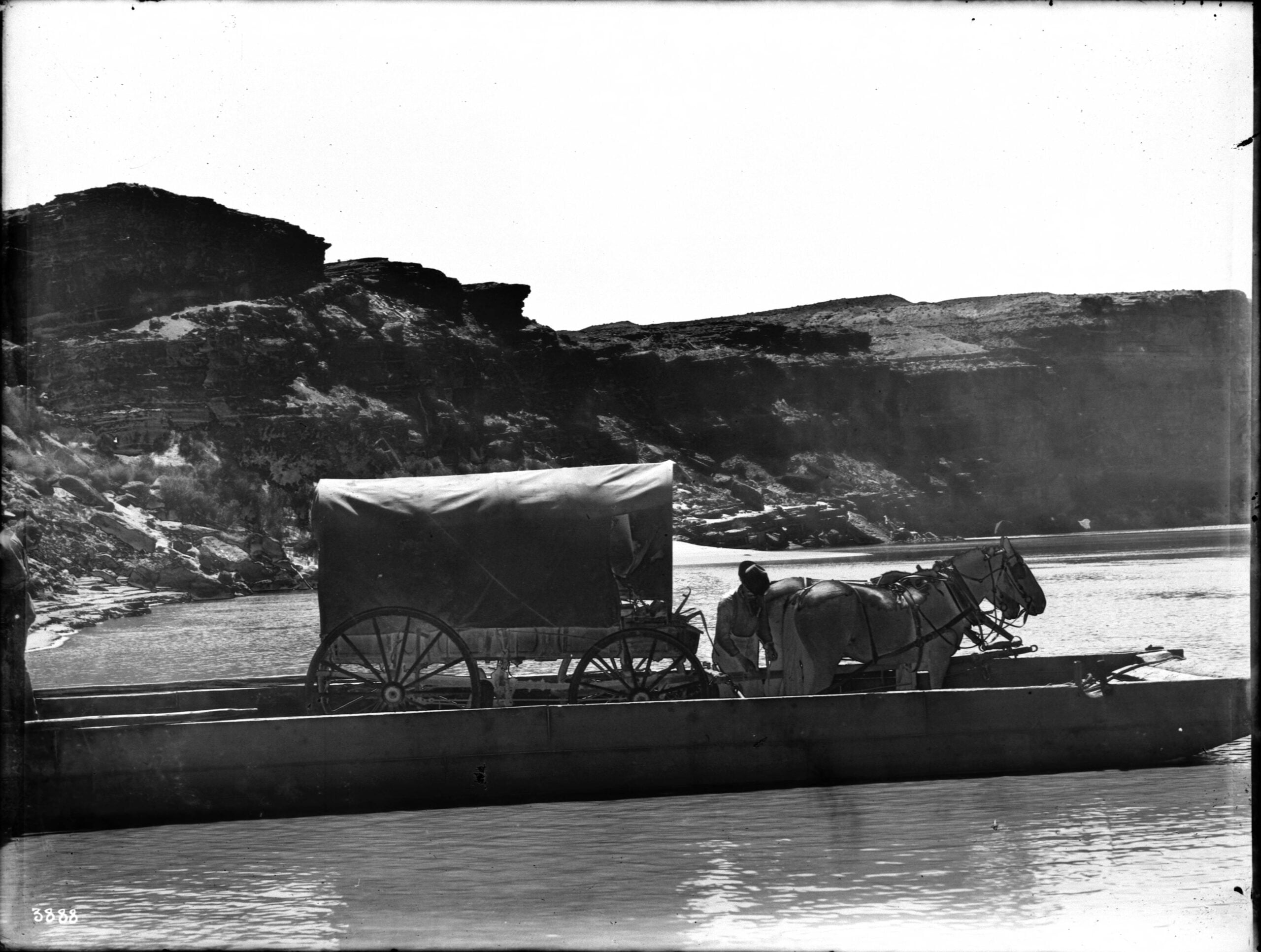 A covered wagon hitched to two horses stands on a wooden ferry crossing a river, with rocky cliffs visible in the background under a bright sky.