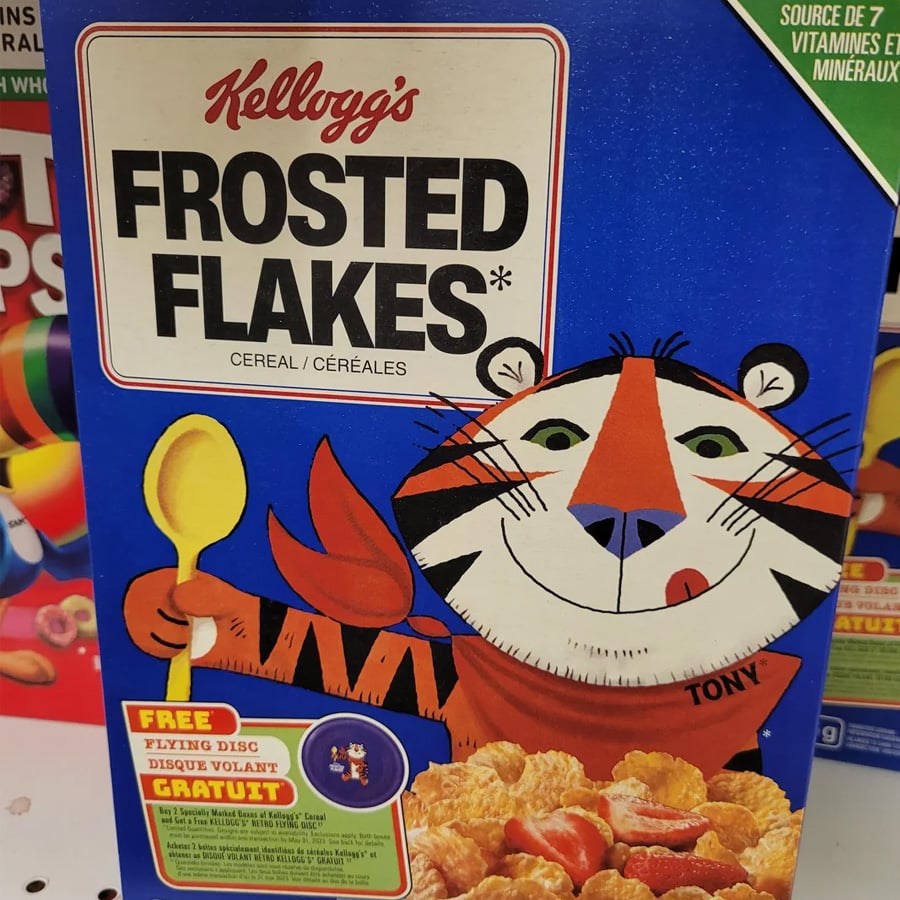 A blue box of Kellogg’s Frosted Flakes cereal featuring Tony the Tiger smiling and holding a yellow spoon. The box offers a free flying disc inside.