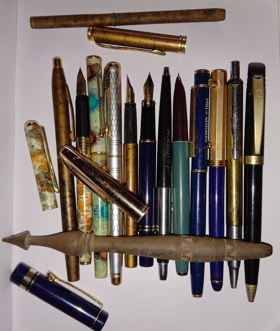 A collection of assorted vintage fountain pens and pen parts, displayed on a white background. The pens have various colors, patterns, and metal finishes, some with their caps removed or set aside.