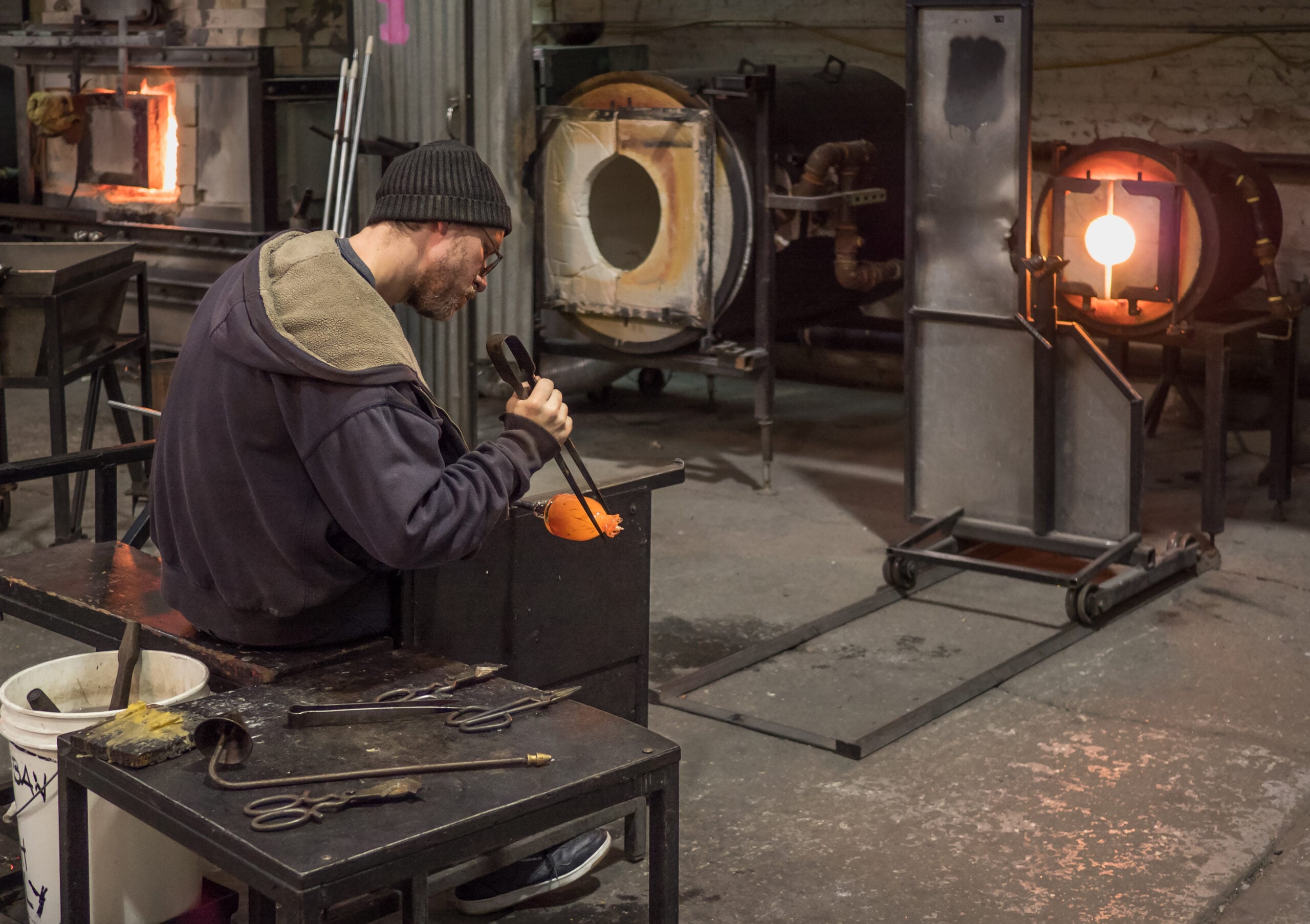 A person wearing a beanie shapes hot glass with tools in a workshop, surrounded by furnaces and equipment, with glowing orange glass visible in the background.