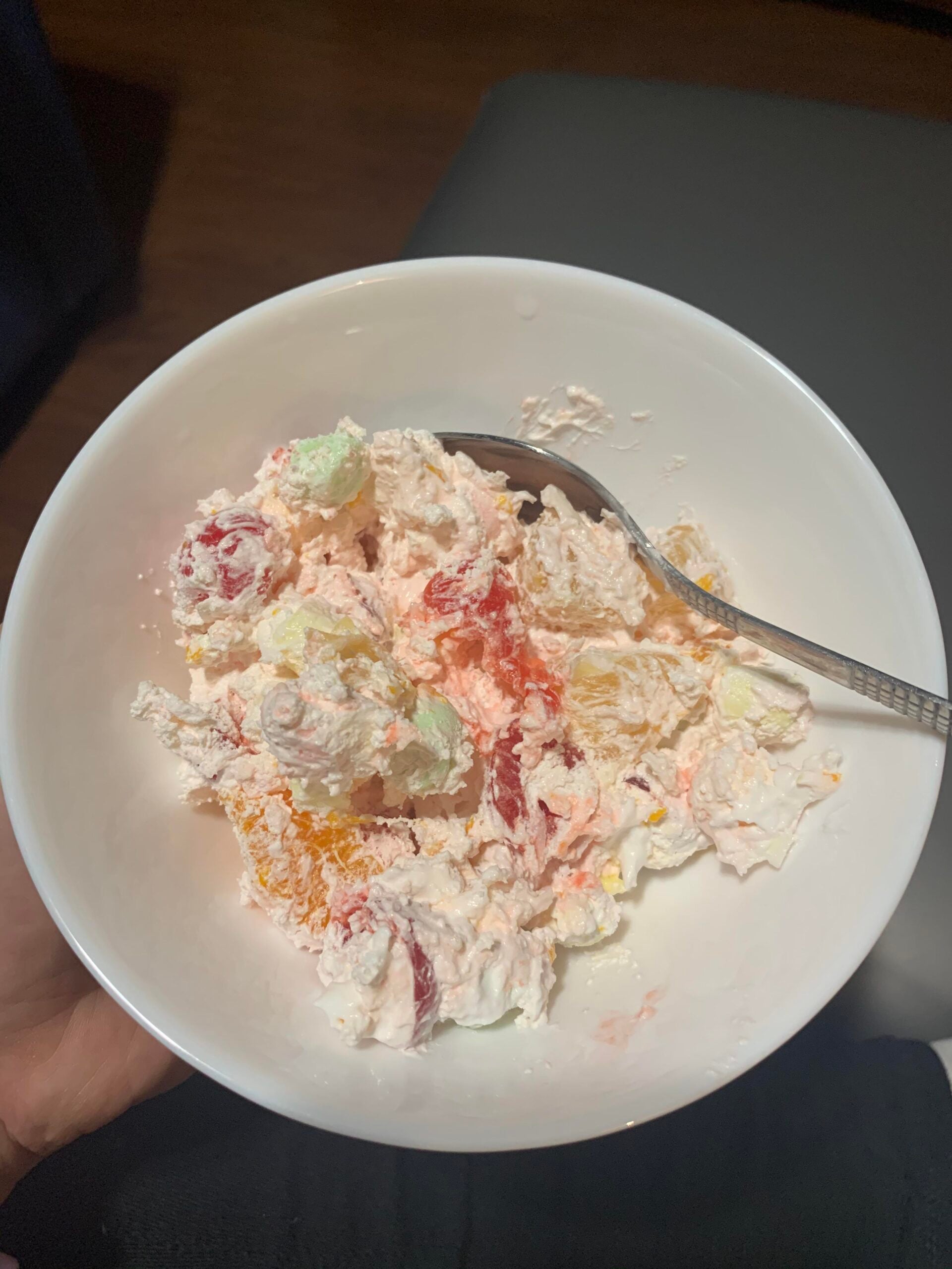 A white bowl filled with fruit salad mixed with whipped cream and colorful marshmallows, with a spoon resting inside.