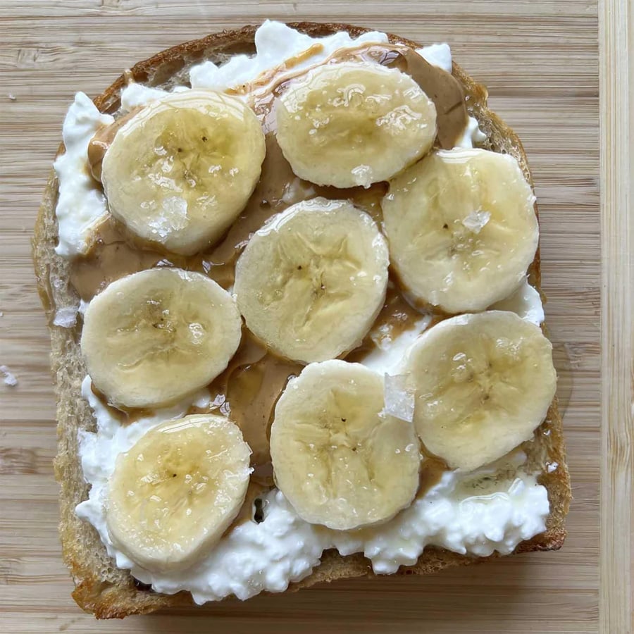 A slice of bread topped with cottage cheese, peanut butter, banana slices, and a drizzle of honey, placed on a wooden surface.