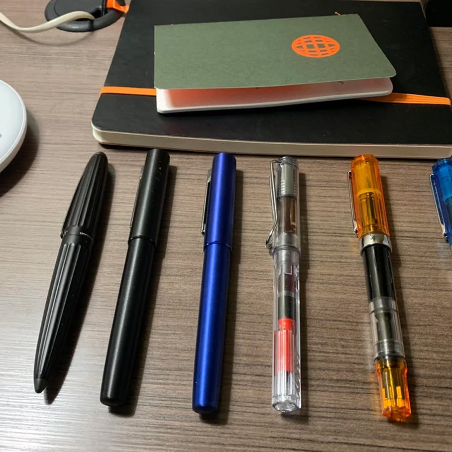 Five fountain pens in different colors and styles are lined up on a wooden desk, with two notebooks and some cords in the background.