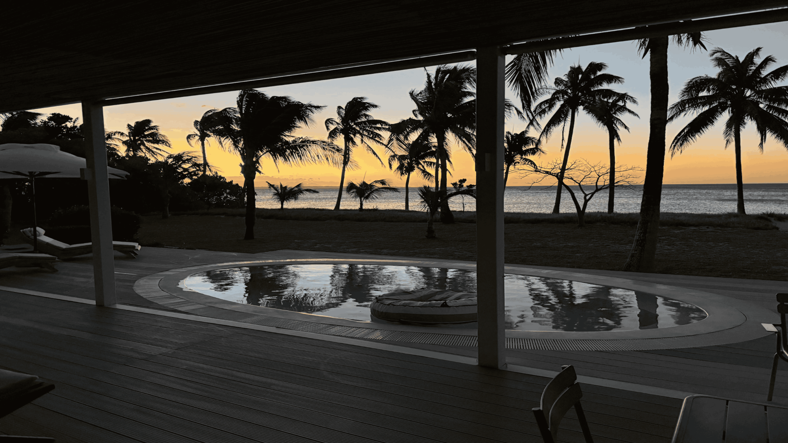 A round pool on a wooden deck overlooks the ocean at sunset, with silhouetted palm trees and lounge chairs creating a tranquil, tropical atmosphere.