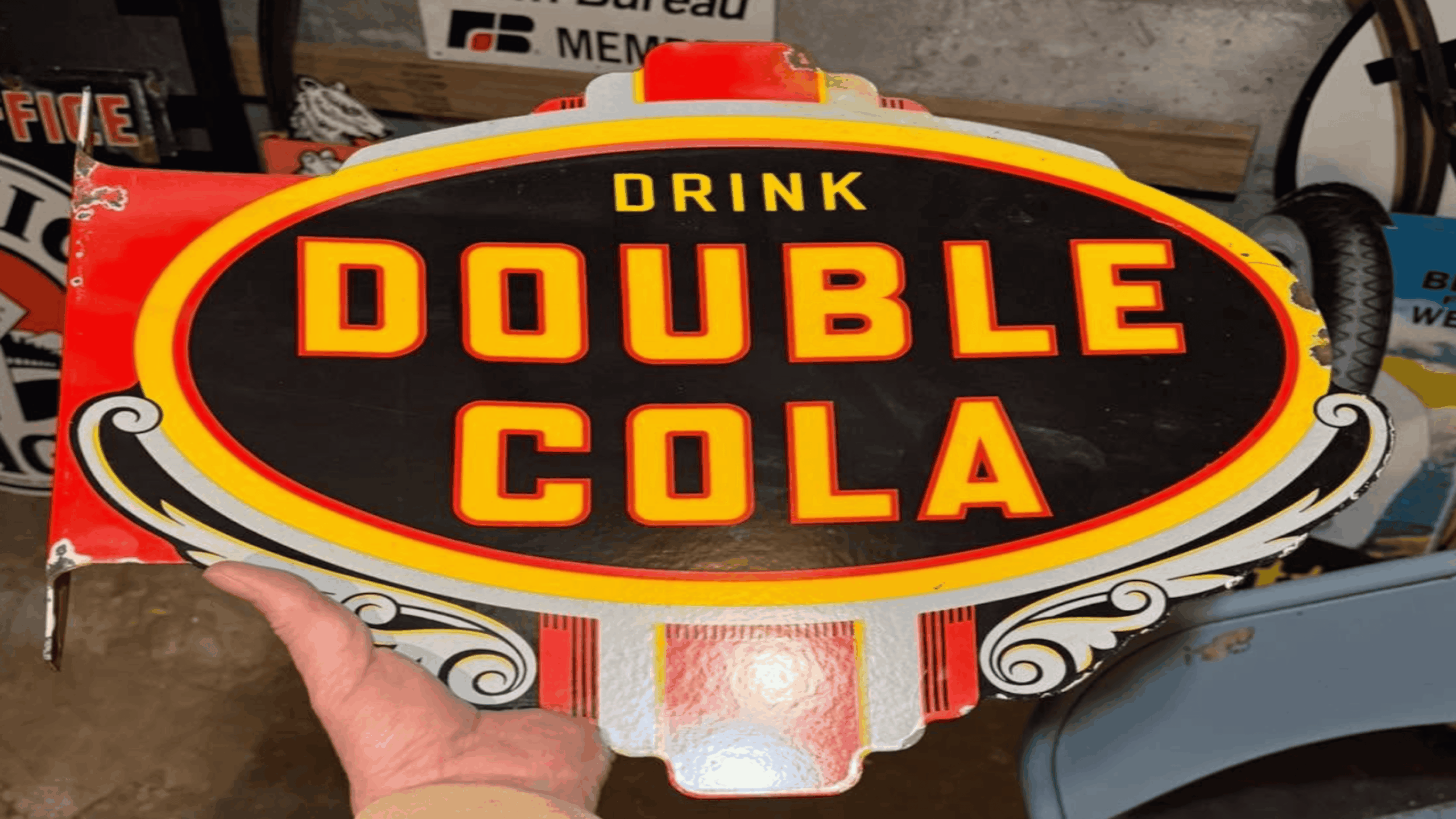 A hand holds a vintage metal sign reading “Drink Double Cola” in bold yellow and red letters on a black oval background with decorative white and red accents.