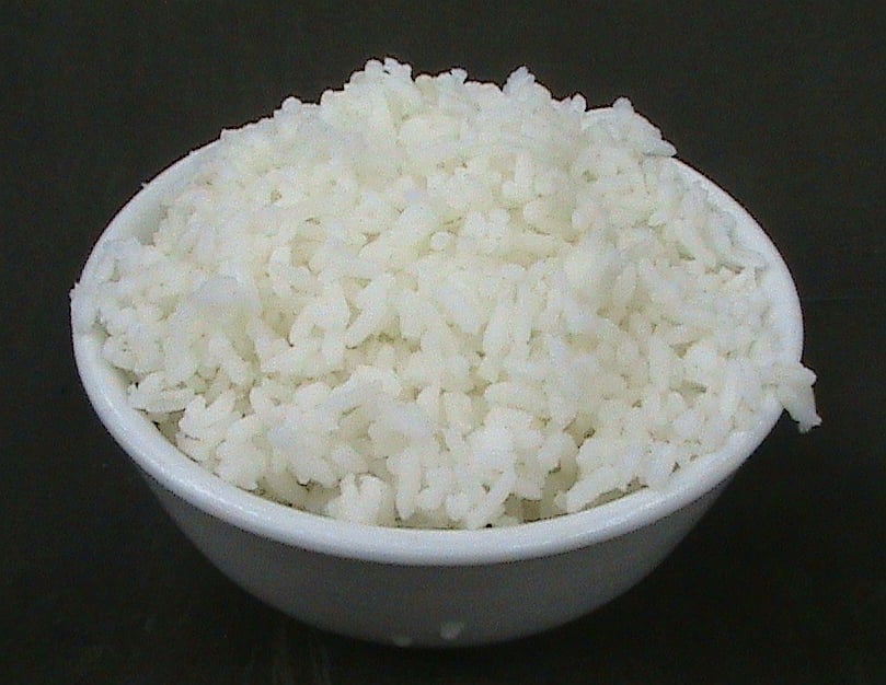 A white ceramic bowl filled with cooked white rice, set against a dark background.