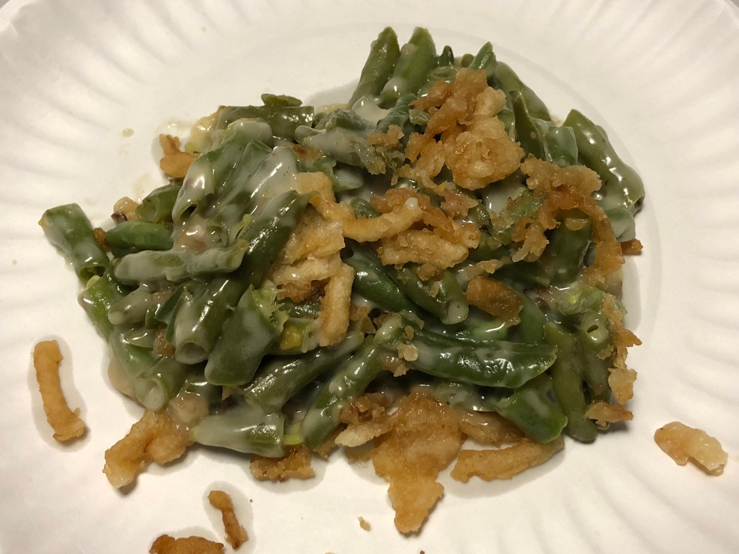 A serving of green bean casserole topped with crispy fried onions on a white paper plate.