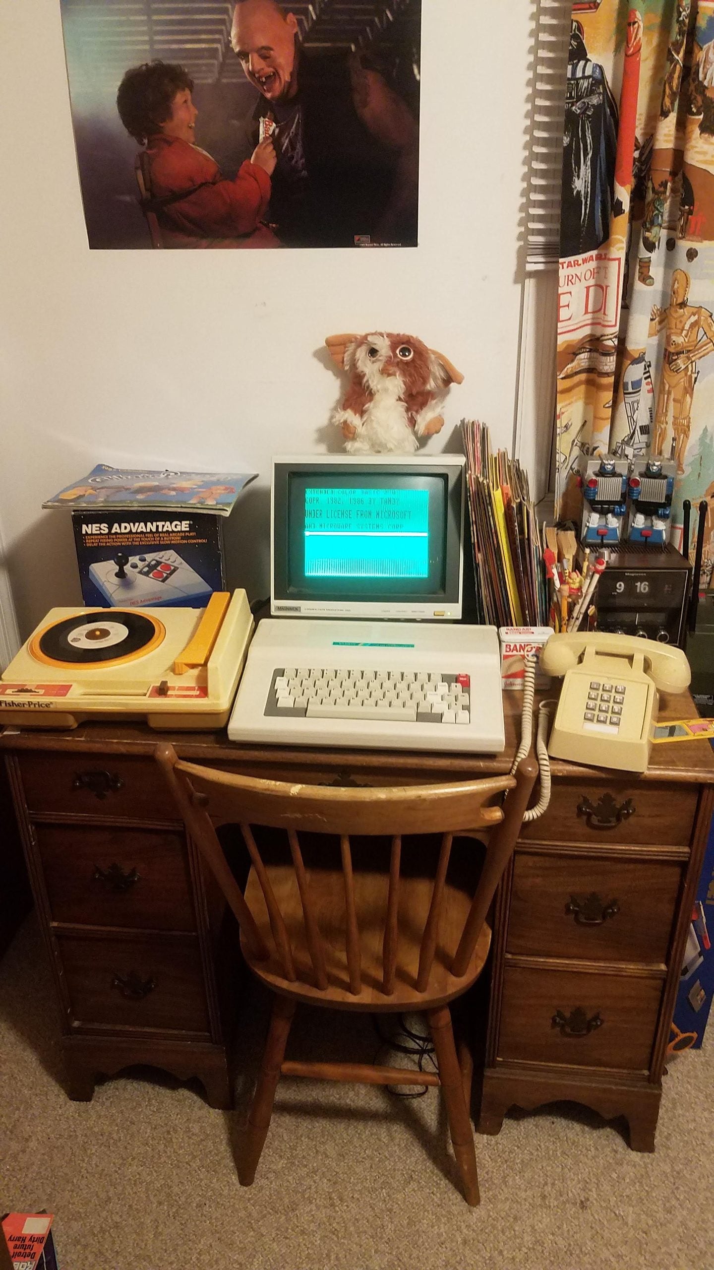 A vintage desk setup with a retro computer, rotary phone, record player, NES Advantage controller, a plush toy, and a poster above. The desk has drawers and a wooden chair is placed in front.