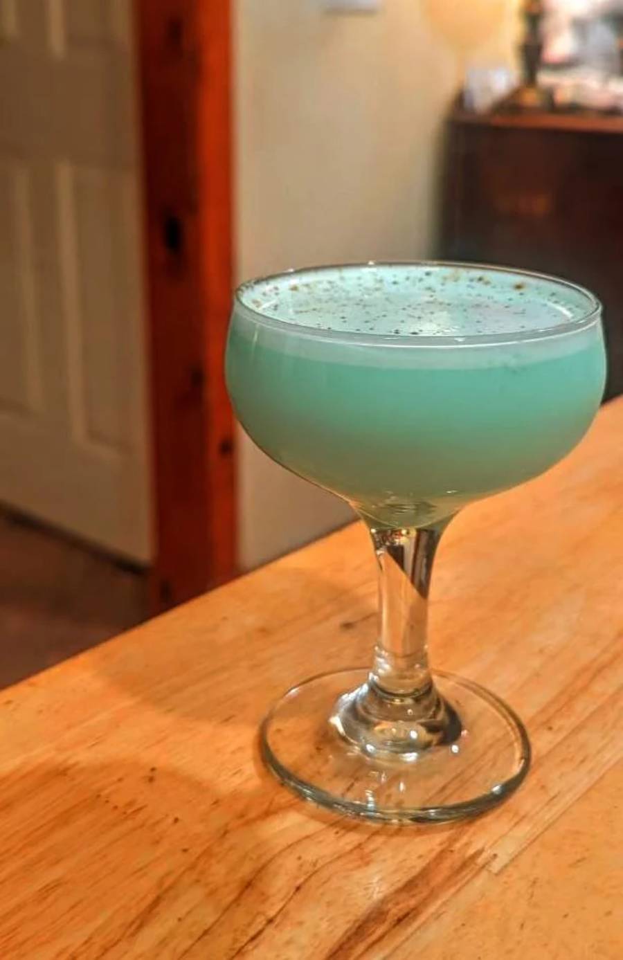 A light blue cocktail in a coupe glass sits on a wooden surface, with a frothy top sprinkled with dark specks. The background is a softly lit interior with a door and some furniture visible.