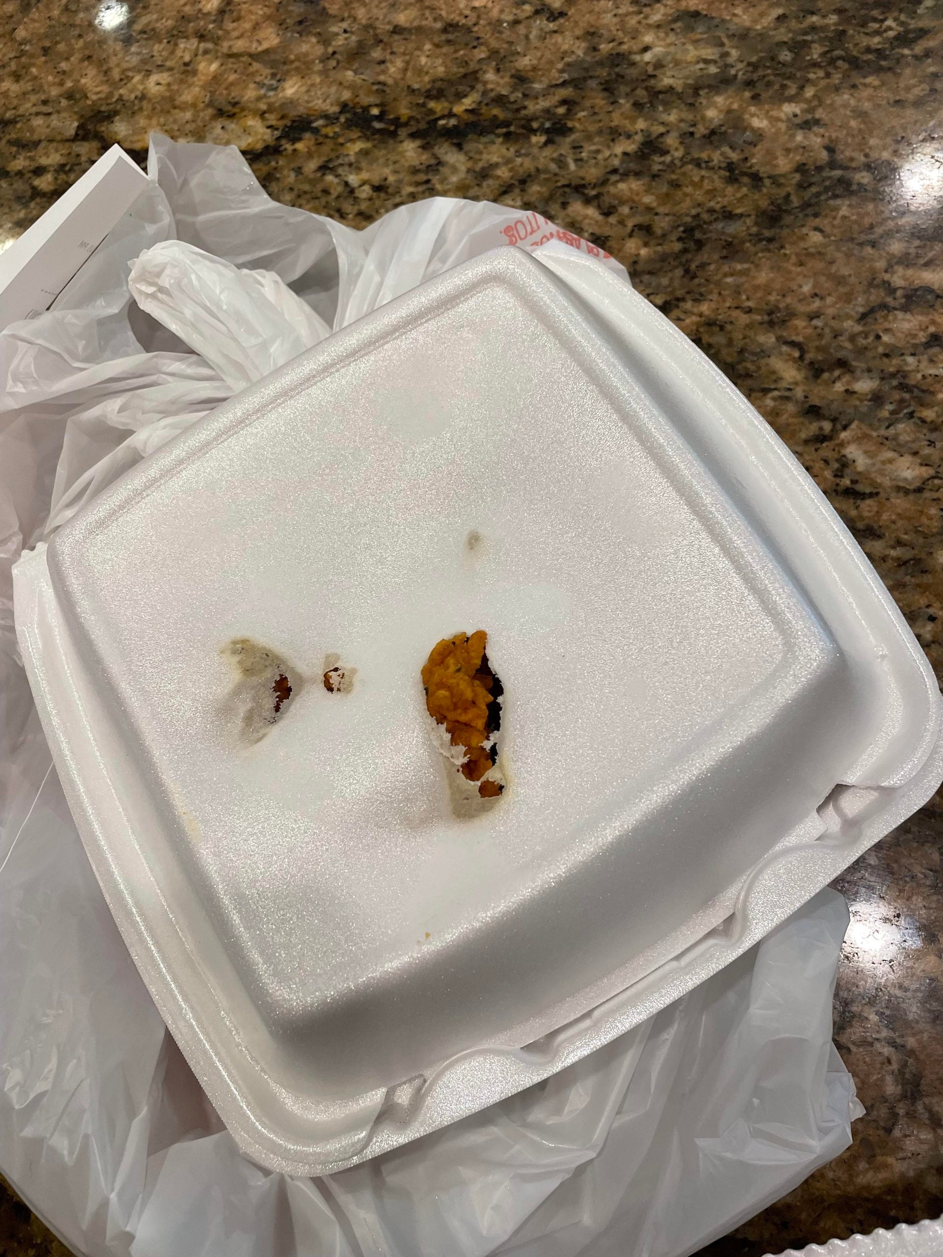 A closed white foam takeout container sits on a plastic bag. There are two small food remnants and a greasy spot on top of the lid. The container is on a brown speckled countertop.