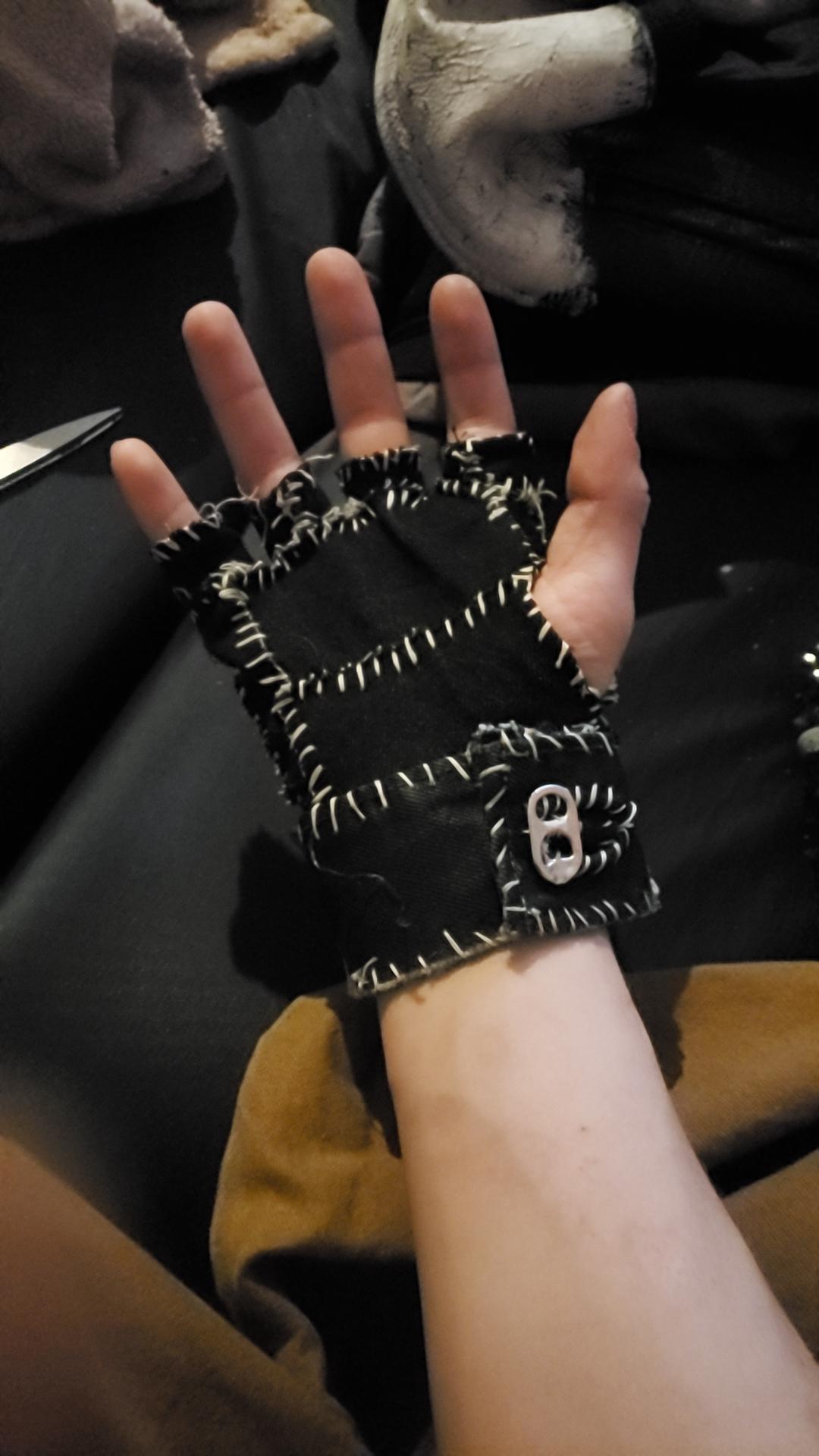 A hand wearing a black, fingerless, patchwork glove with white stitching and a small metal buckle on the wrist. Scissors and fabric are visible in the background.
