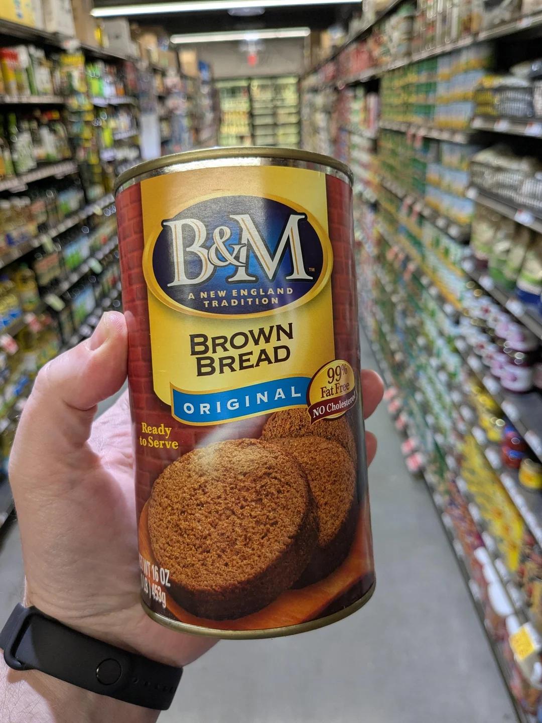 A hand holds a can of B&M Brown Bread Original in a grocery store aisle filled with various products on shelves.