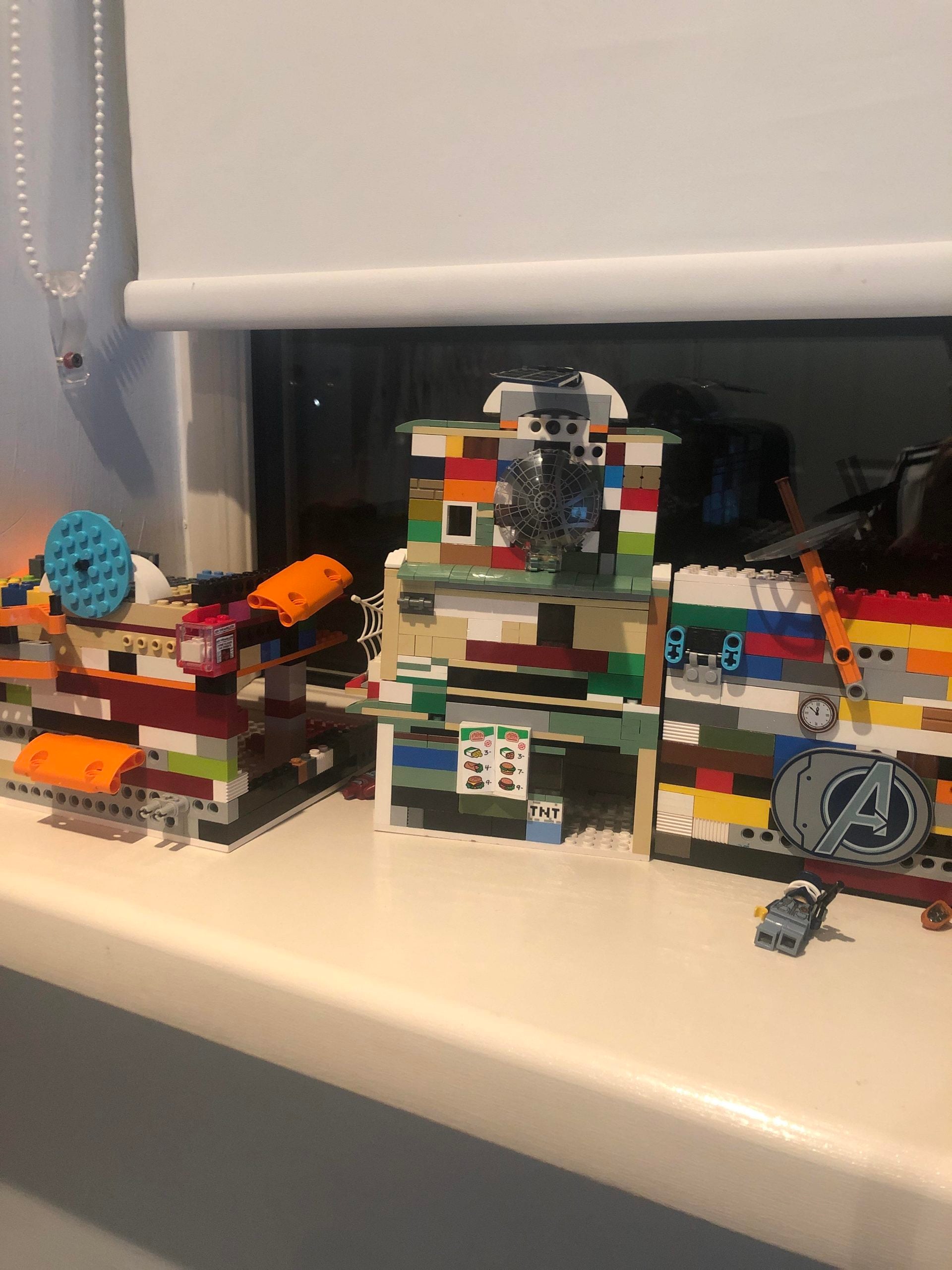 Three colorful LEGO structures sit on a windowsill, each built from assorted bricks. One features the Avengers logo, another has a TNT sticker, and small LEGO accessories are scattered nearby.