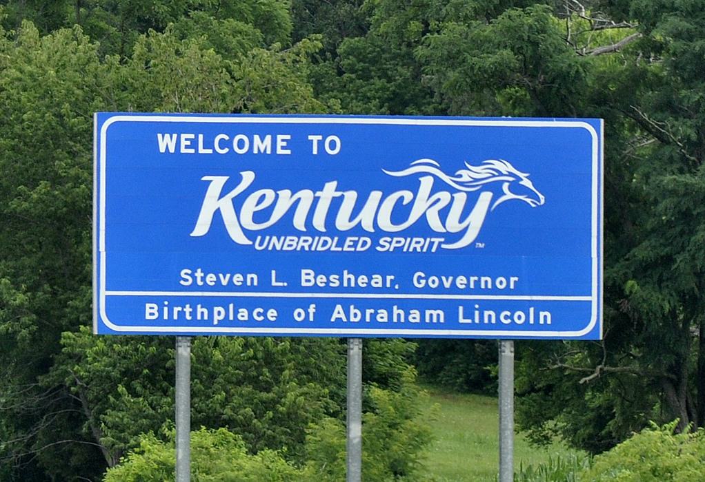 A blue road sign reads: “Welcome to Kentucky, Unbridled Spirit. Steven L. Beshear, Governor. Birthplace of Abraham Lincoln.” Trees and greenery are in the background.