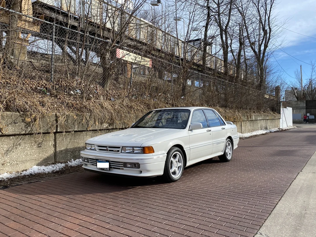 A white four-door sedan is parked on a brick road next to a concrete wall and bare trees, with an elevated train track visible in the background.