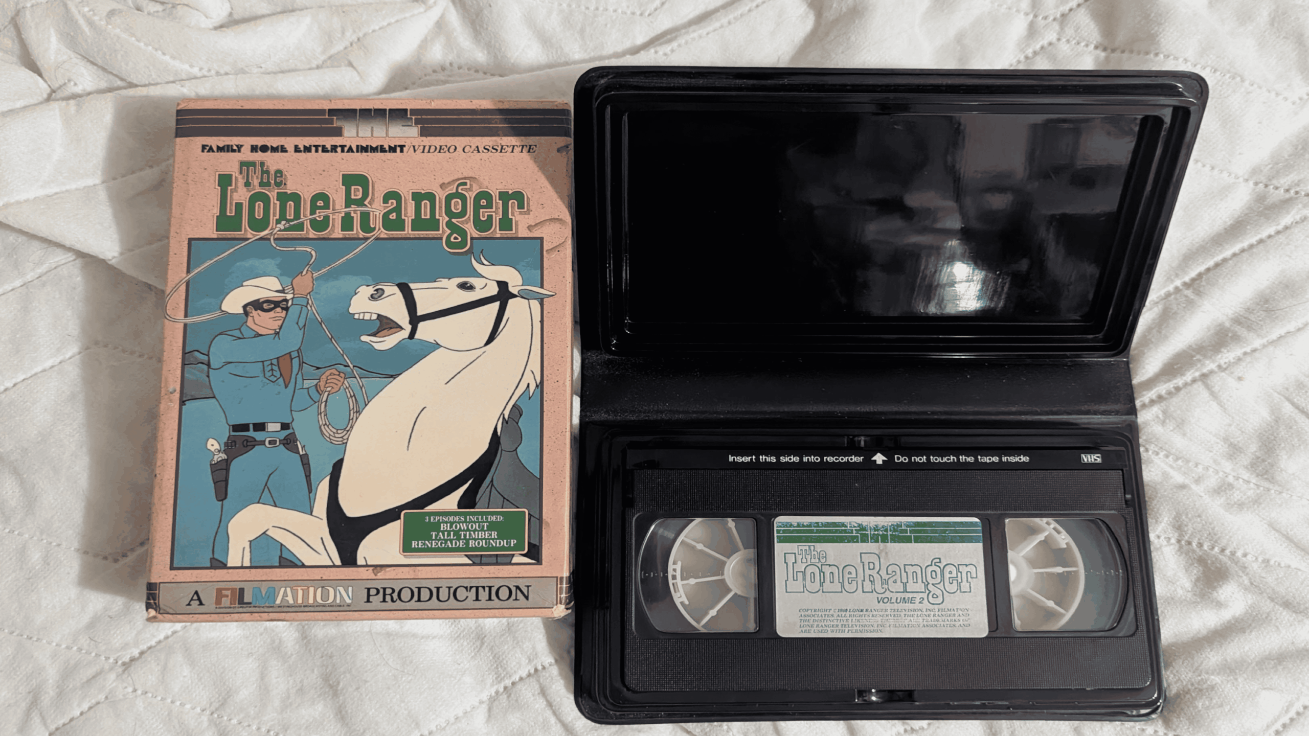 A VHS tape labeled “The Lone Ranger Vol. 1” sits in an open black case next to its illustrated cardboard sleeve, featuring the Lone Ranger and his horse on a cream-colored quilted surface.