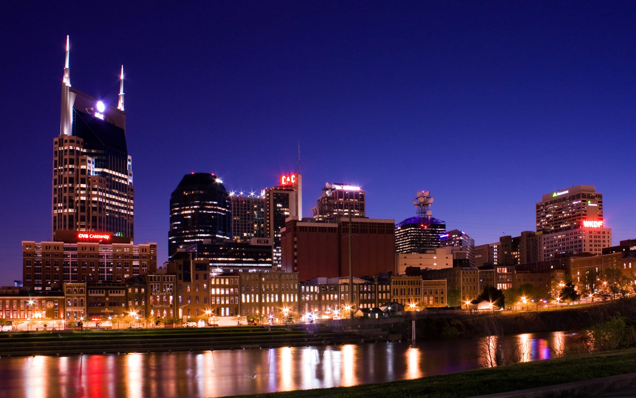 Nashville city skyline at night with illuminated buildings reflecting on the Cumberland River under a deep blue sky.
