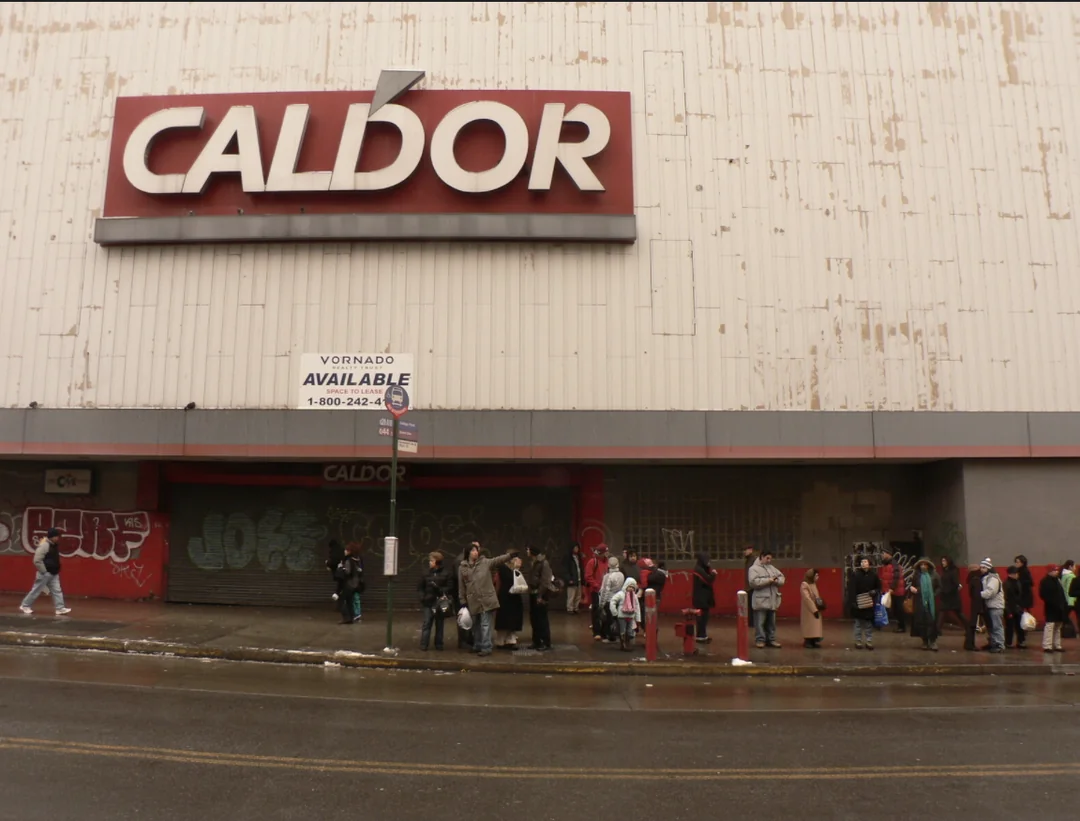A group of people stand in line outside a weathered, graffiti-covered Caldor storefront with a large red and white sign above them. The building appears old and closed.