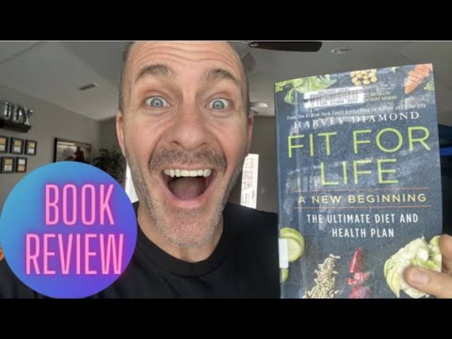 A smiling man holds up the book “Fit For Life” by Harvey Diamond. Next to him is a circular graphic with the text “BOOK REVIEW.” Indoor background with pictures on the wall.