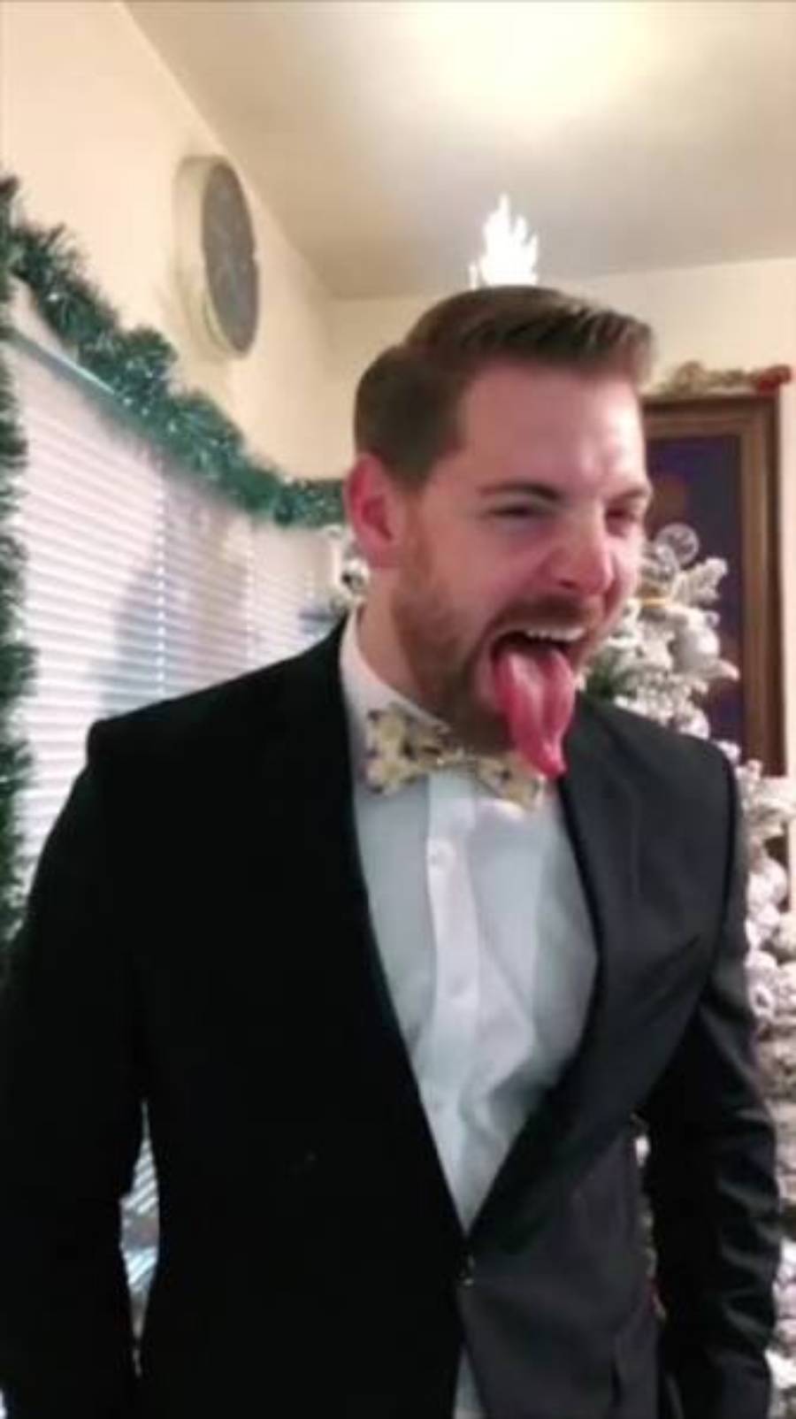 A man in a suit and floral bow tie stands indoors, sticking his tongue out playfully. There is a decorated Christmas tree and holiday garland in the background.