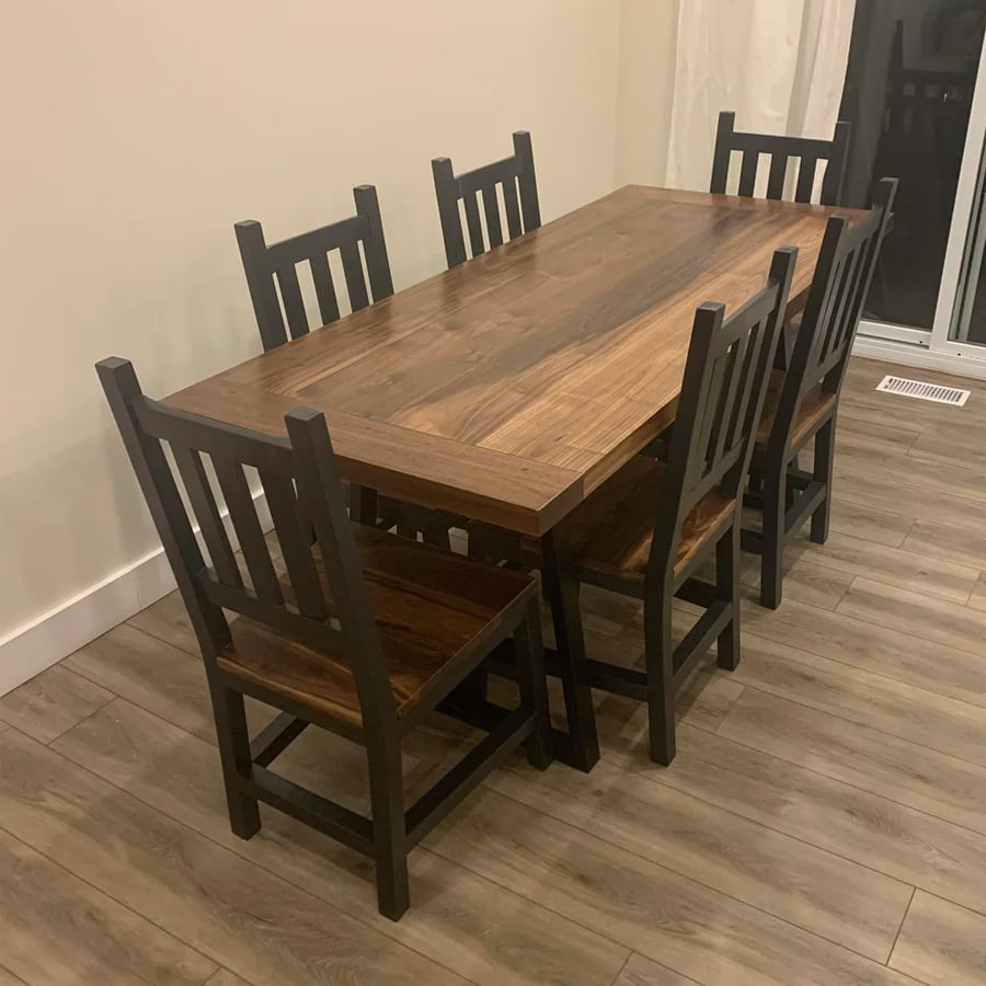 A wooden dining table with six matching chairs is arranged in a room with wood flooring and light-colored walls. The table and chairs have a dark frame with natural wood tops and seats.