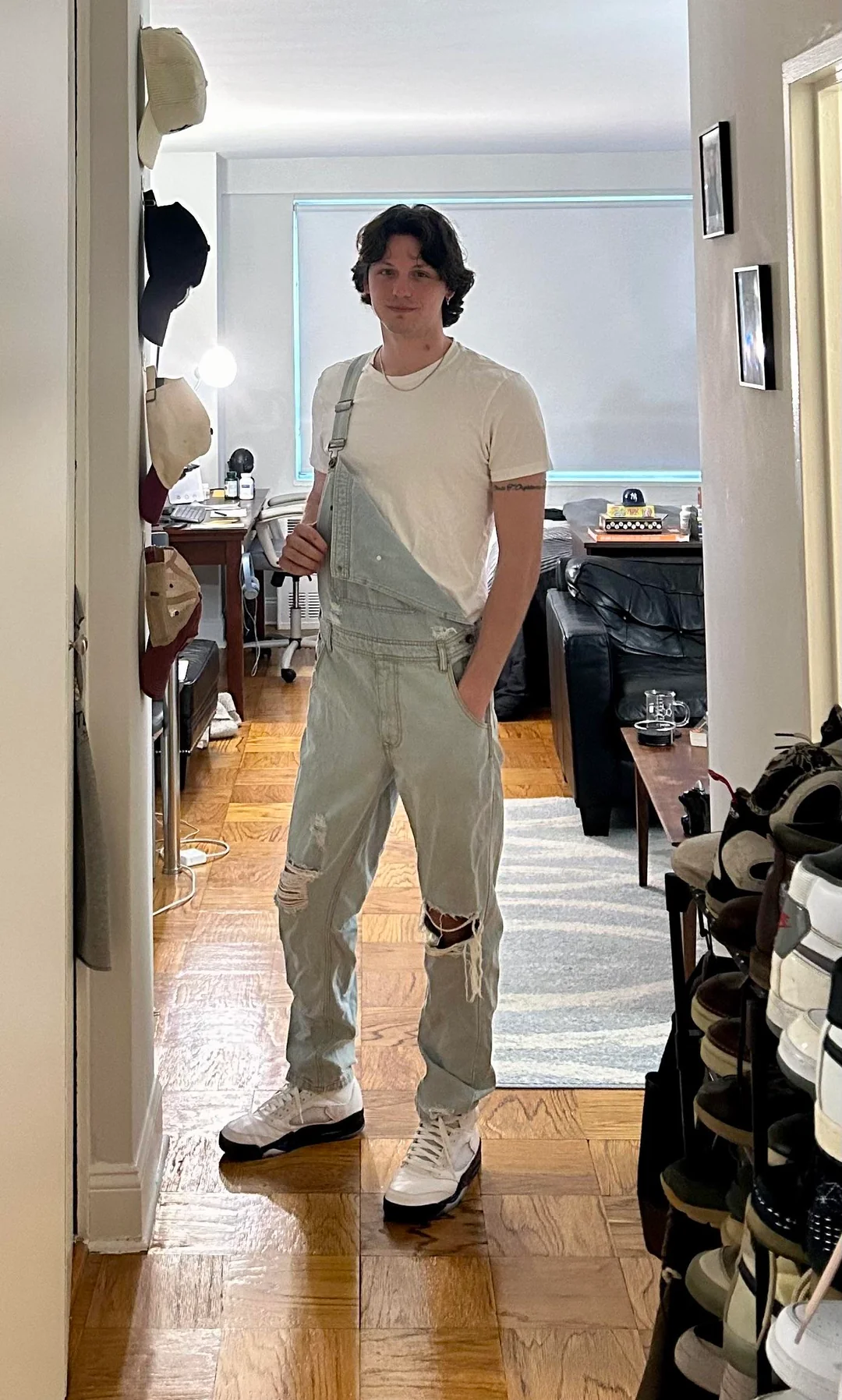 A person stands indoors wearing light blue overalls with one strap undone, a white t-shirt, and white sneakers. They are in a hallway with hats on the wall and shoes on shelves, with a living room visible in the background.