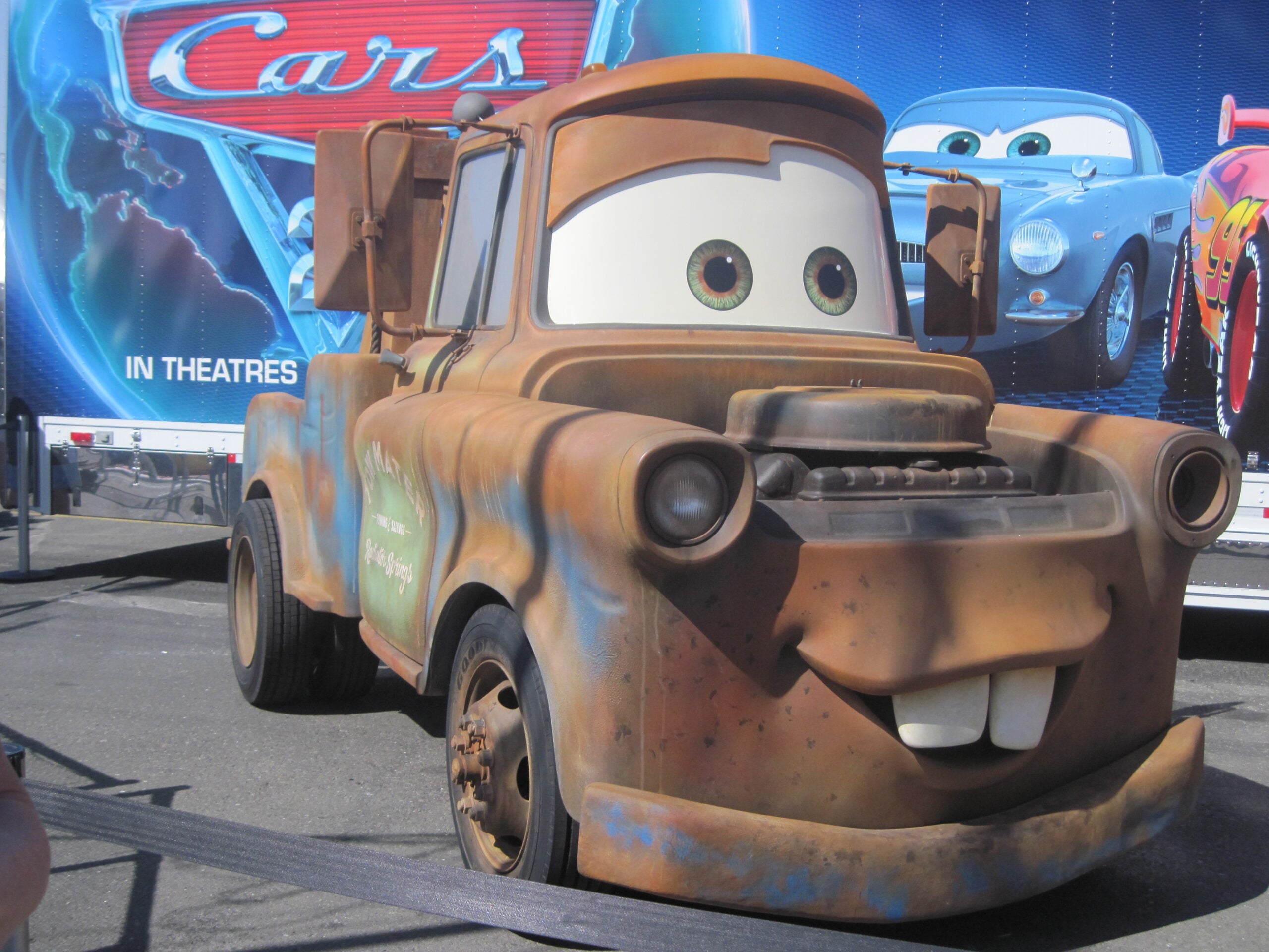 Rusty brown tow truck character from the animated movie “Cars,” smiling in front of a blue “Cars” movie poster backdrop, with other car characters visible in the background.