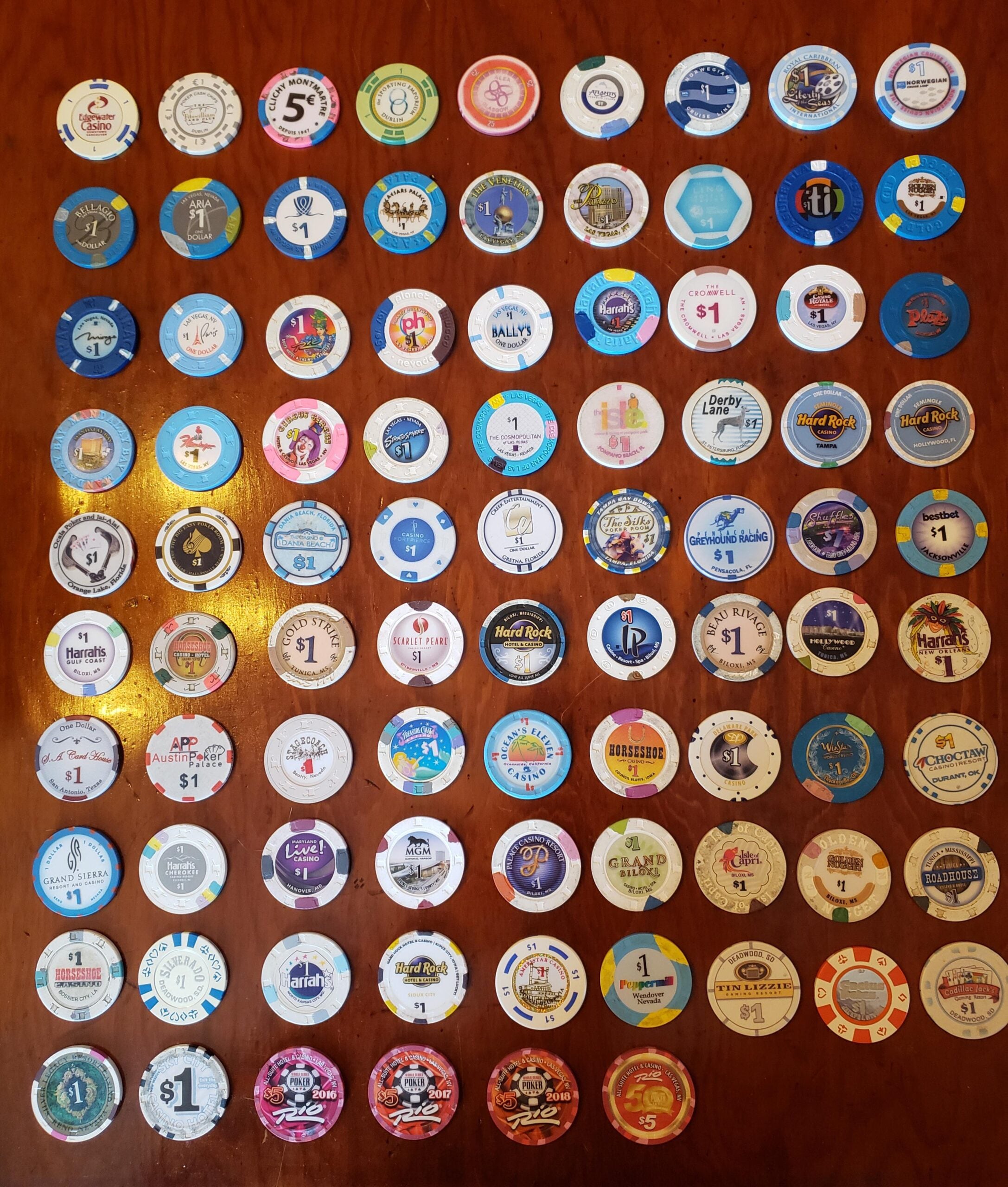 Collection of colorful $1 casino chips from various casinos, arranged in neat rows on a wooden surface. Each chip features unique logos, designs, and colors representing different establishments.