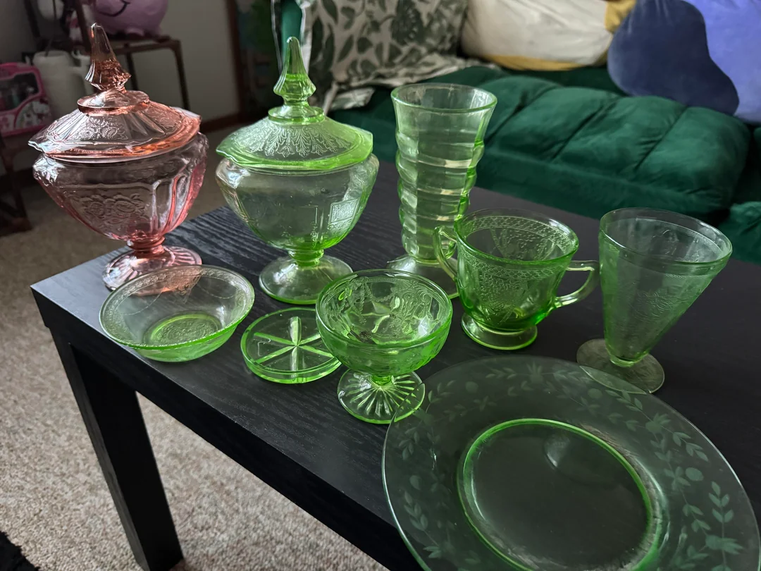 A variety of vintage glassware, mostly green with one pink piece, is displayed on a black table in a living room with a green couch and decorative pillows in the background.