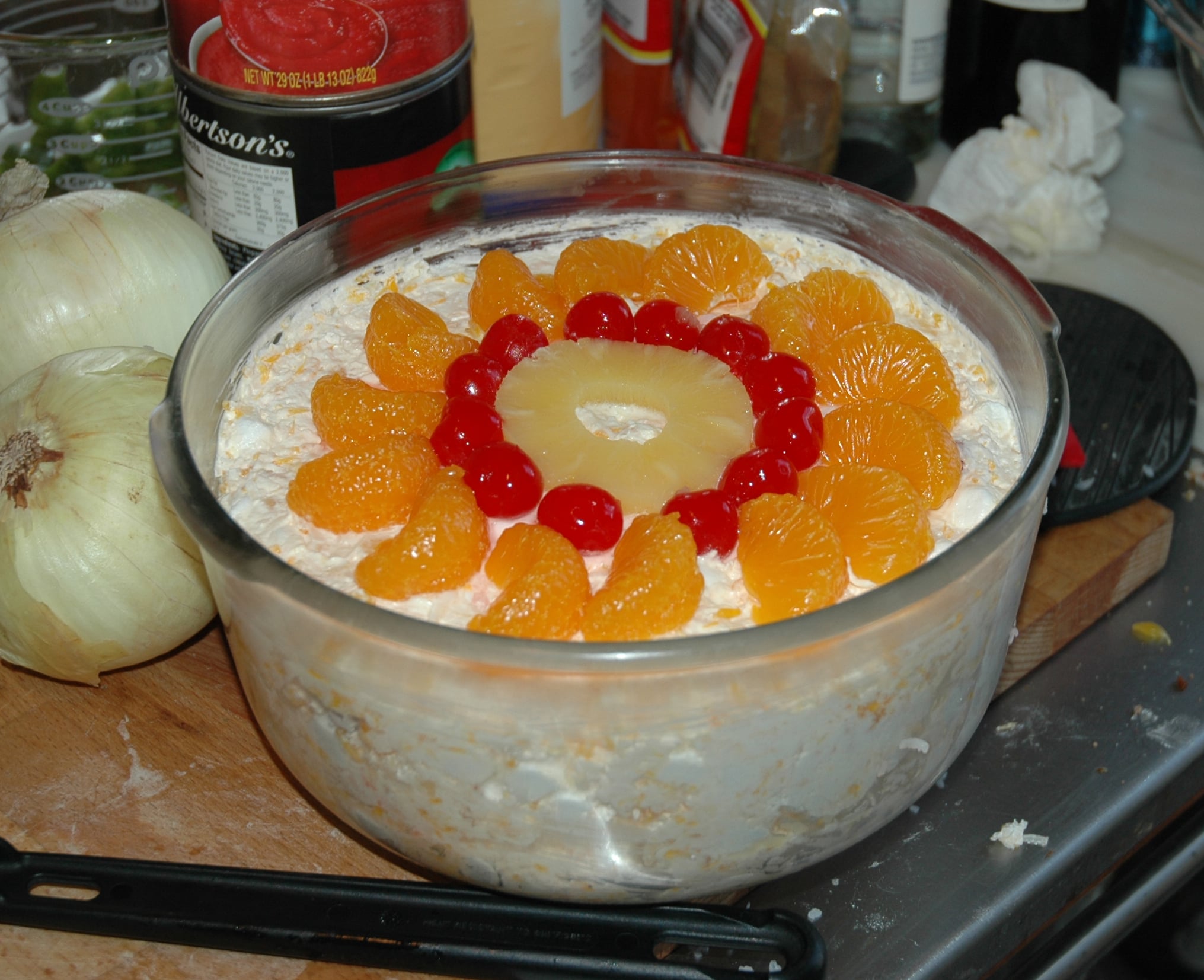 A glass bowl of ambrosia salad topped with a ring of orange segments, cherries, and a pineapple slice, on a kitchen counter with onions and various items nearby.