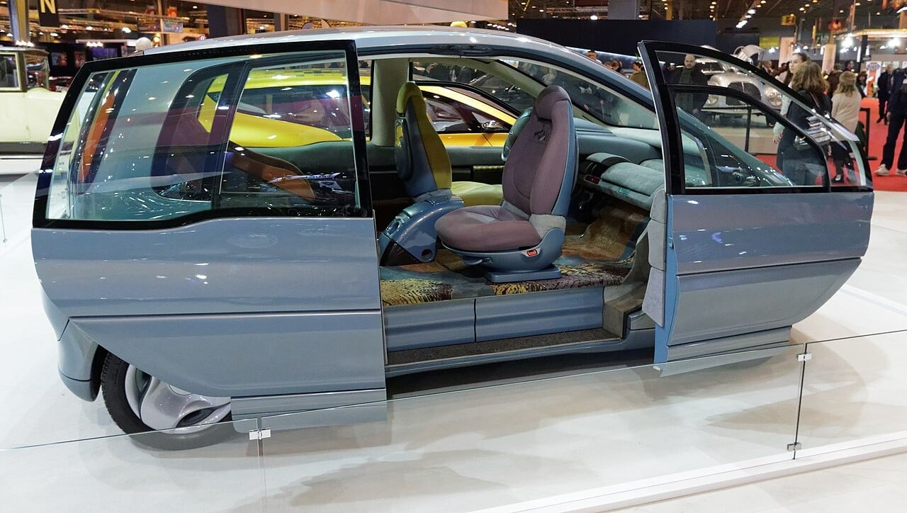 A futuristic blue concept car with large windows and sliding doors is displayed at an auto show, revealing its spacious interior and unique seating arrangement.