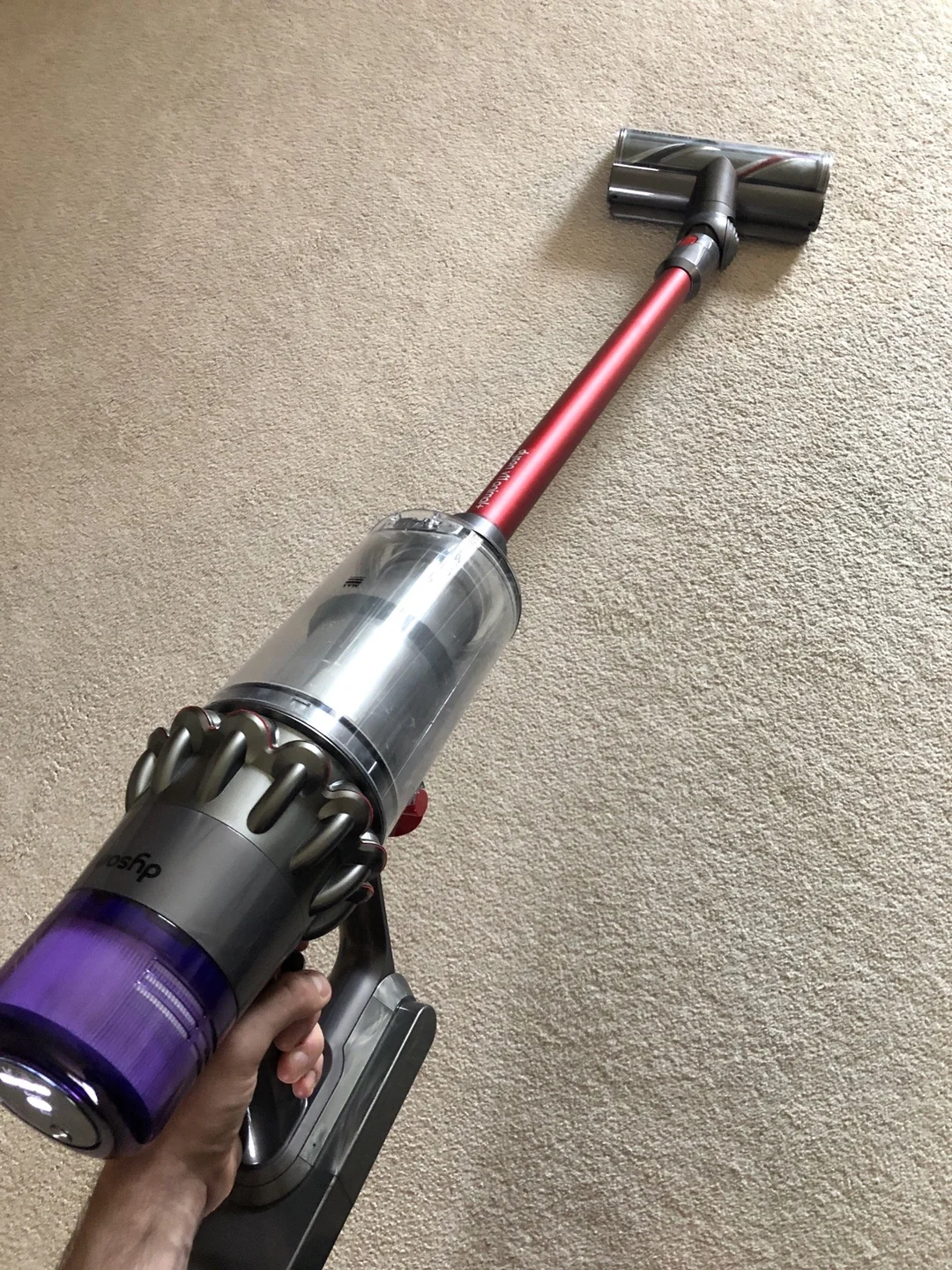 A person holds a cordless stick vacuum cleaner with a red and silver handle, cleaning a beige carpet.