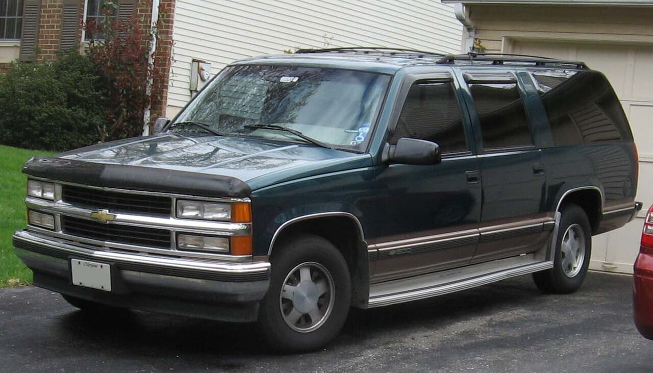 A dark green Chevrolet Suburban SUV is parked in a driveway next to a house with white siding and brick accents. The vehicle is facing left and has a roof rack and silver trim.