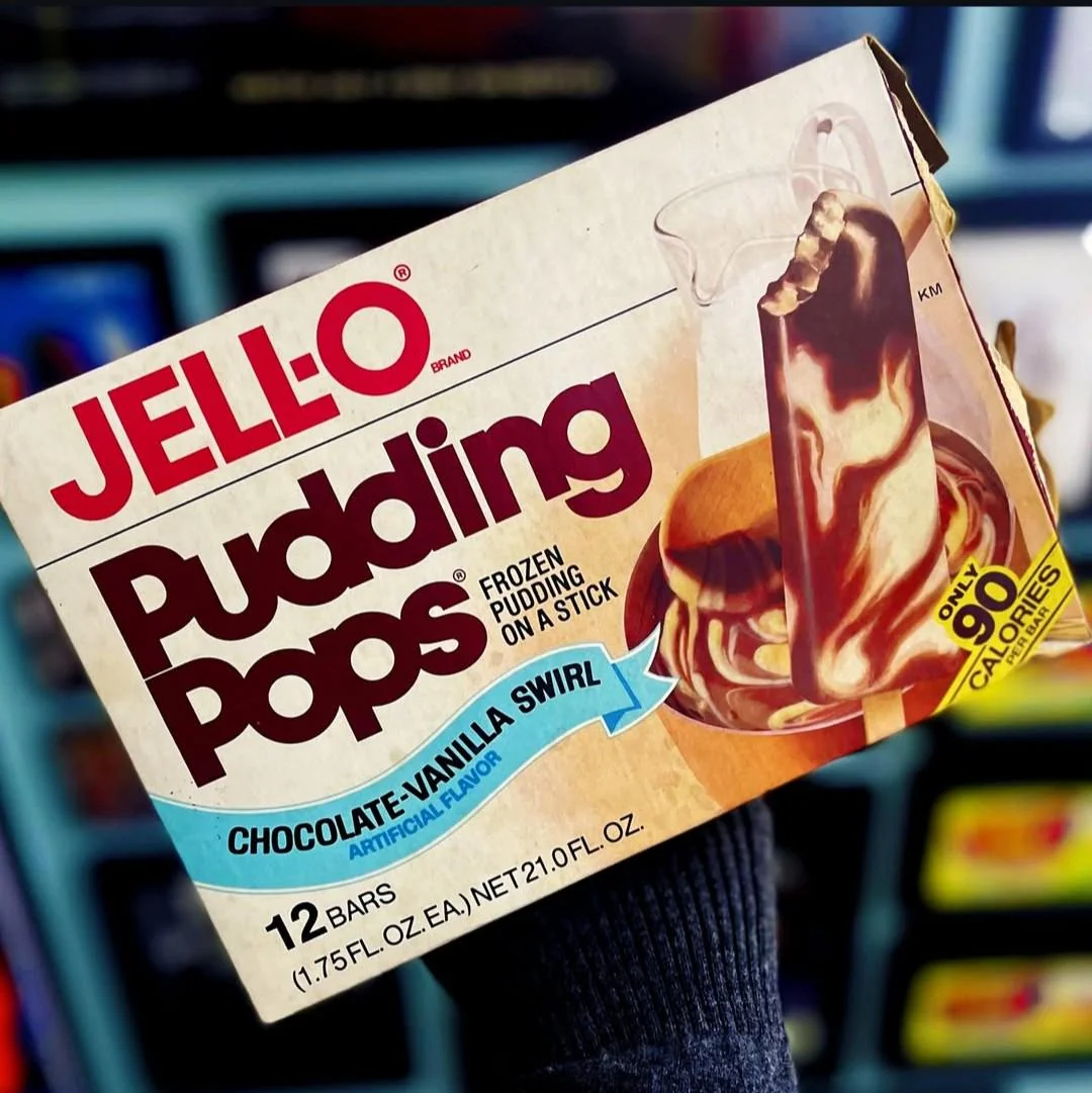 A hand holds a box of Jell-O Pudding Pops, chocolate-vanilla swirl flavor. The box shows an image of the frozen pudding treat and notes it contains 12 bars, each under 90 calories.