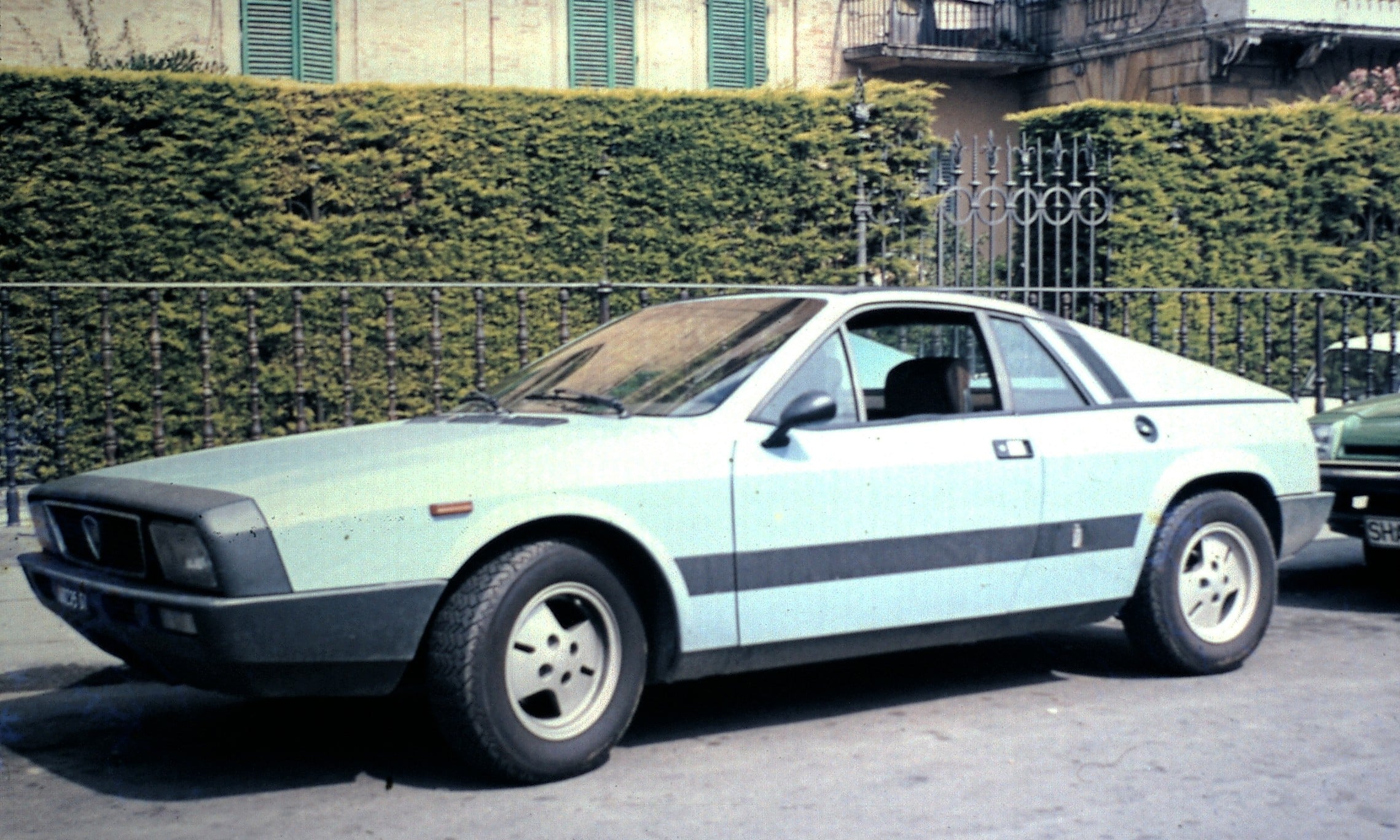 A light blue vintage sports car is parked on a street in front of a hedge and wrought iron fence, with another car partially visible behind it. The background includes a yellow building with green window shutters.