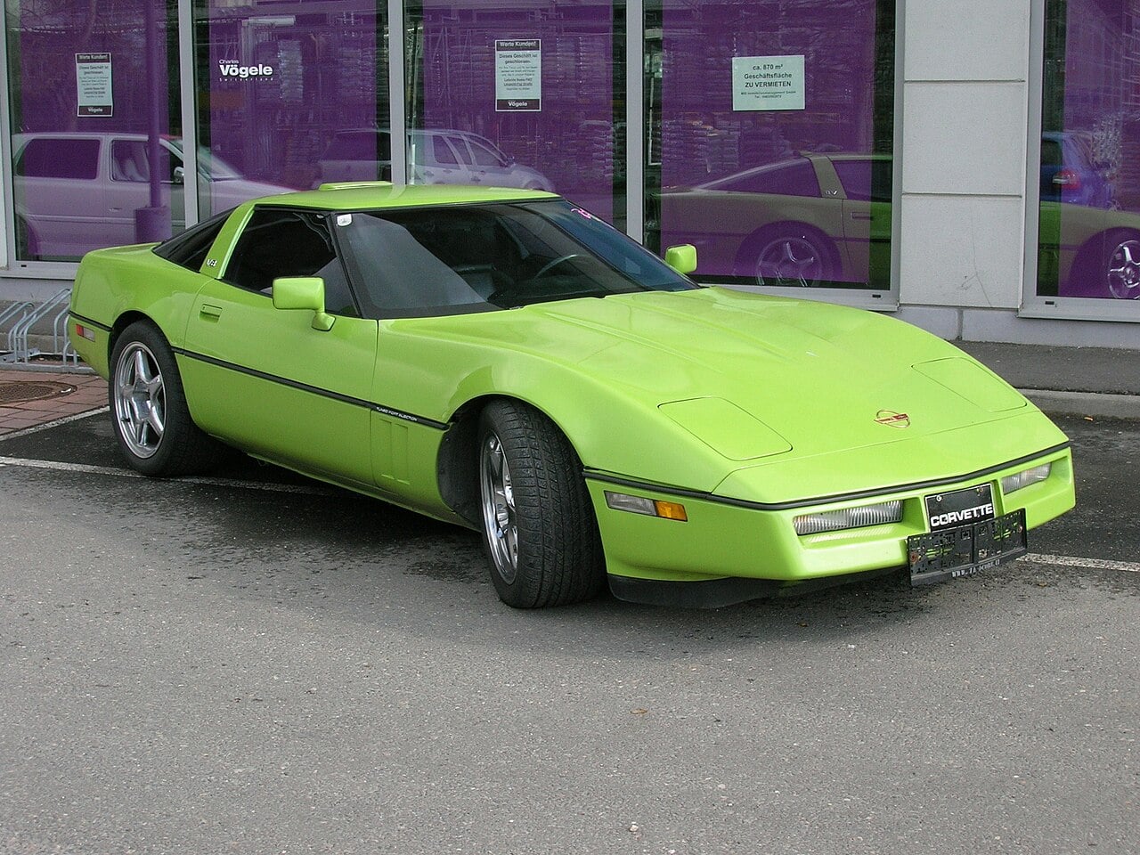 A bright green sports car is parked in front of large purple-tinted windows, with its front wheels turned outward and a reflection of the car visible in the glass behind it.