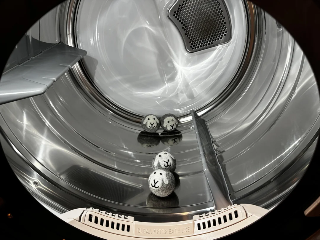 Four dryer balls with cute sheep faces rest inside a shiny, empty metal dryer drum, viewed from the door opening. A label at the bottom reads “CLEAN AFTER EACH USE.”
