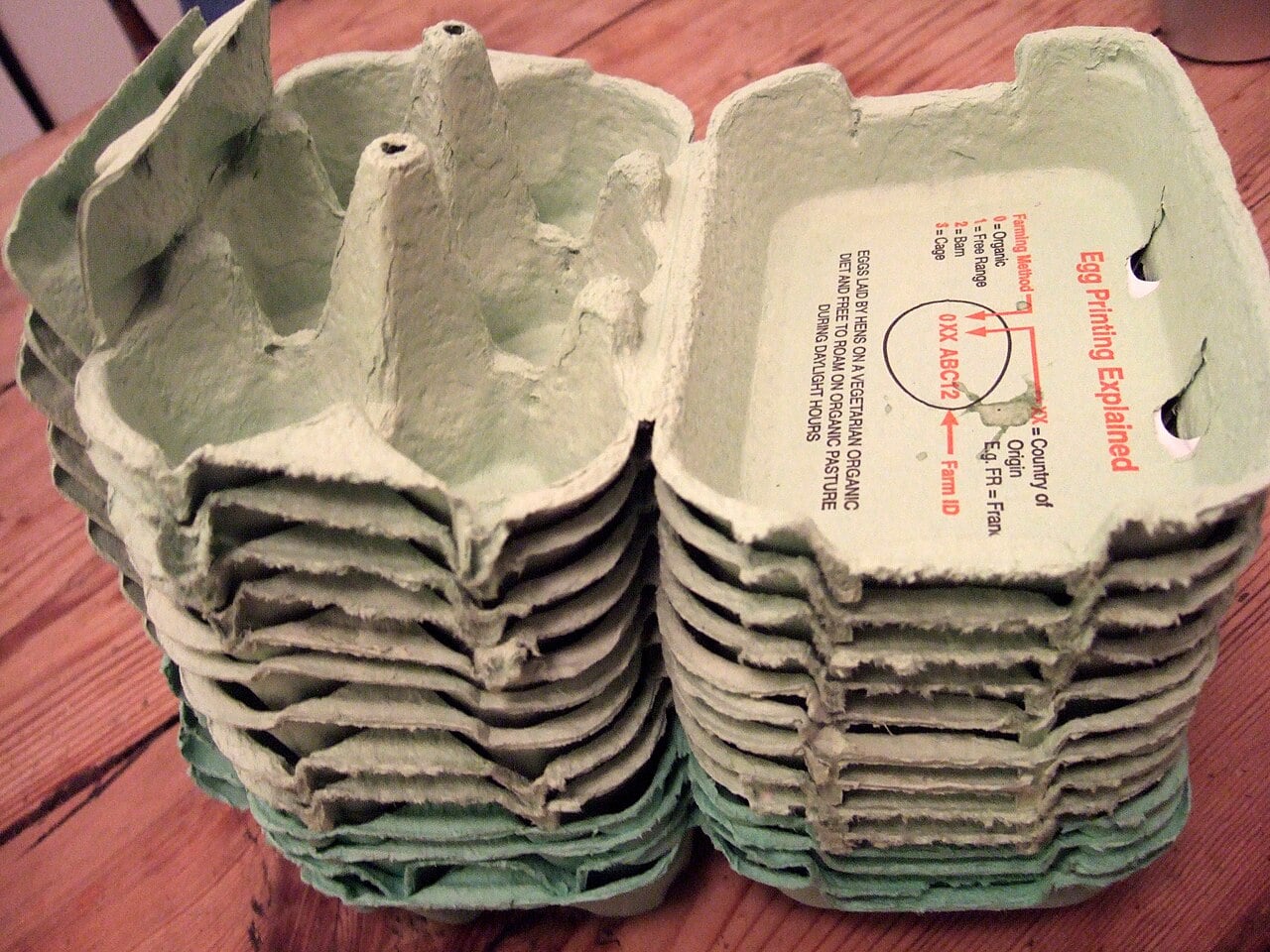 A stack of empty, green, cardboard egg cartons sits on a wooden surface. The carton on top has red printed text and a diagram explaining egg printing.