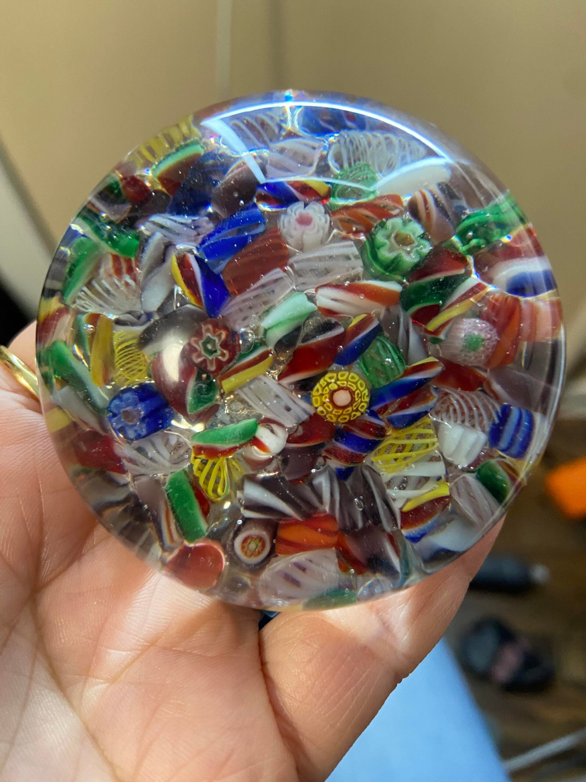 A hand holds a round glass paperweight filled with colorful, intricate millefiori glass patterns against a blurred indoor background.