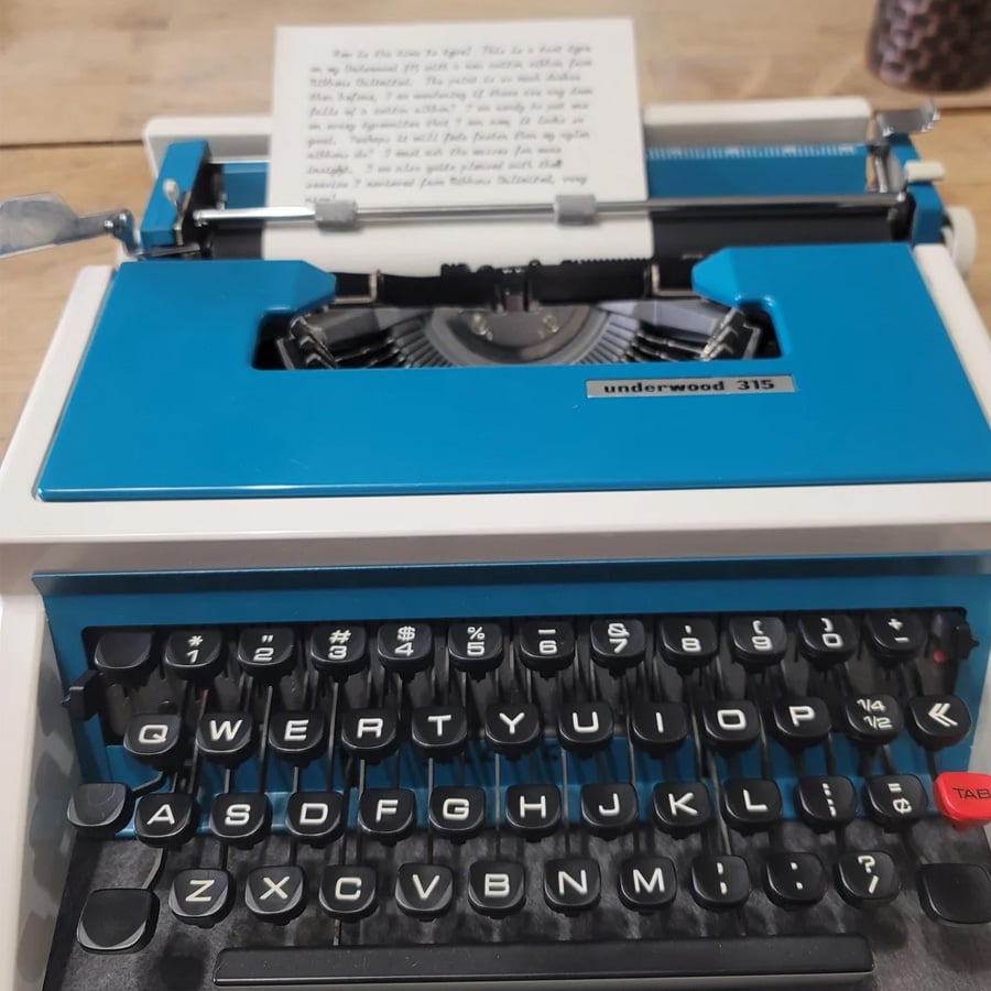 A blue and white Underwood 315 typewriter with a piece of paper loaded, sitting on a wooden table. The keys are visible in the foreground.