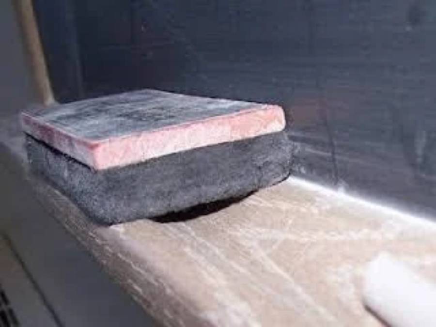A close-up of a chalkboard eraser resting on a wooden chalk tray, with a piece of white chalk nearby and chalk dust visible on the tray and eraser.