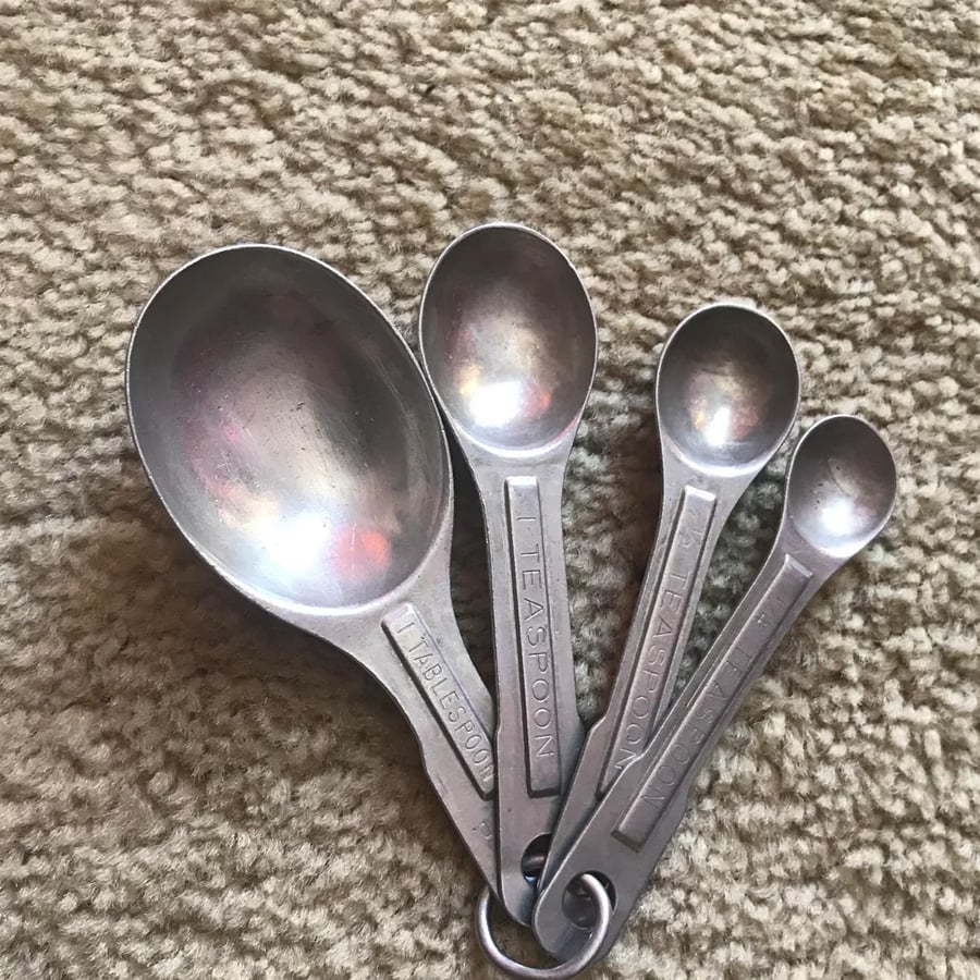 Four metal measuring spoons of different sizes, labeled tablespoon and teaspoon, are nested together on a metal ring and placed on a textured beige carpet.
