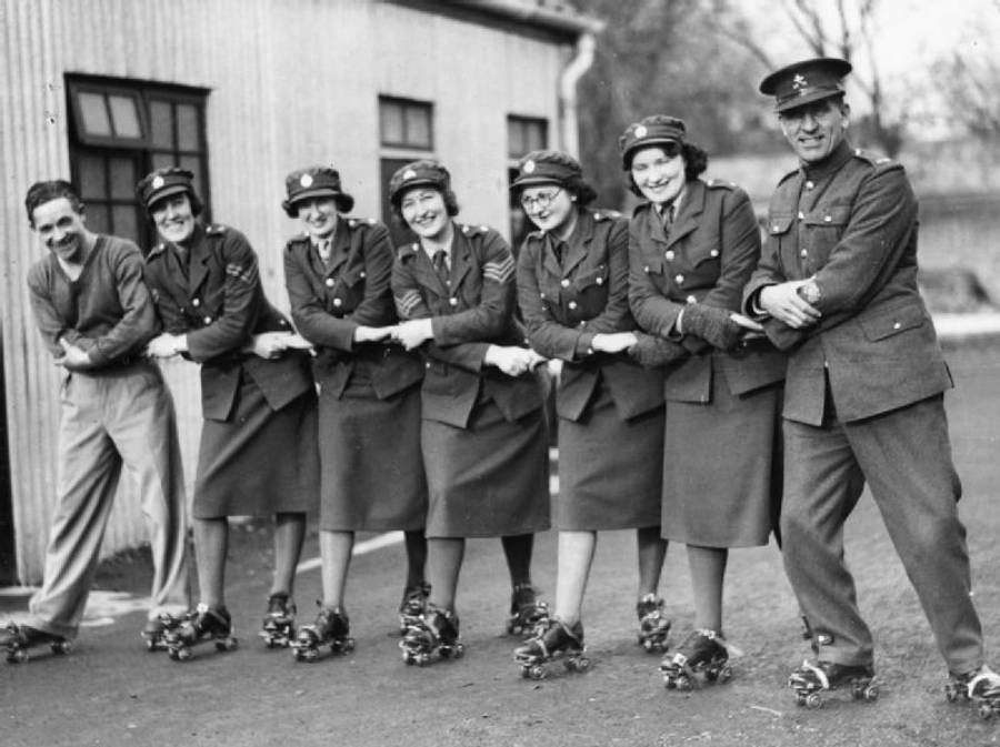 Six women in uniform and one man stand outside, smiling and linking arms while posing on roller skates. The group appears to be enjoying themselves and are dressed in mid-20th-century attire.