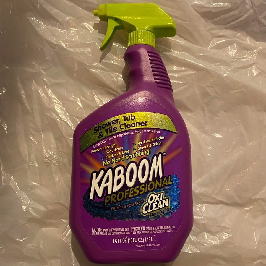 A purple spray bottle of Kaboom Professional Shower, Tub & Tile Cleaner with OxiClean, placed on a crumpled plastic surface. The bottle has a bright green spray nozzle and yellow label accents.