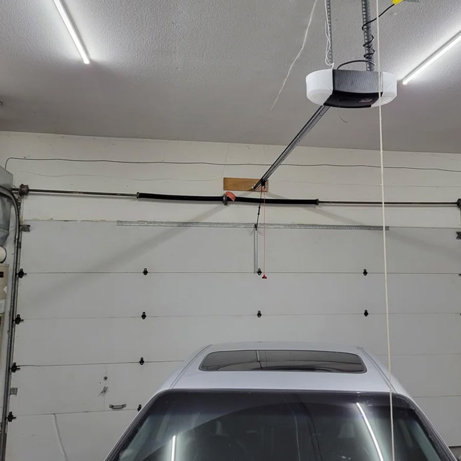 Garage interior with a car parked beneath a partially visible garage door. A garage door opener and a white rope hang from the ceiling, and fluorescent lights illuminate the space.