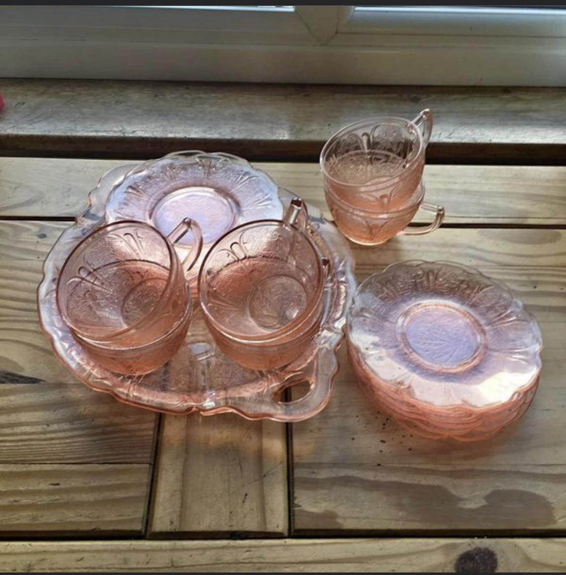A set of translucent pink vintage glass dishware with cups, saucers, and plates arranged on a wooden table near a window.