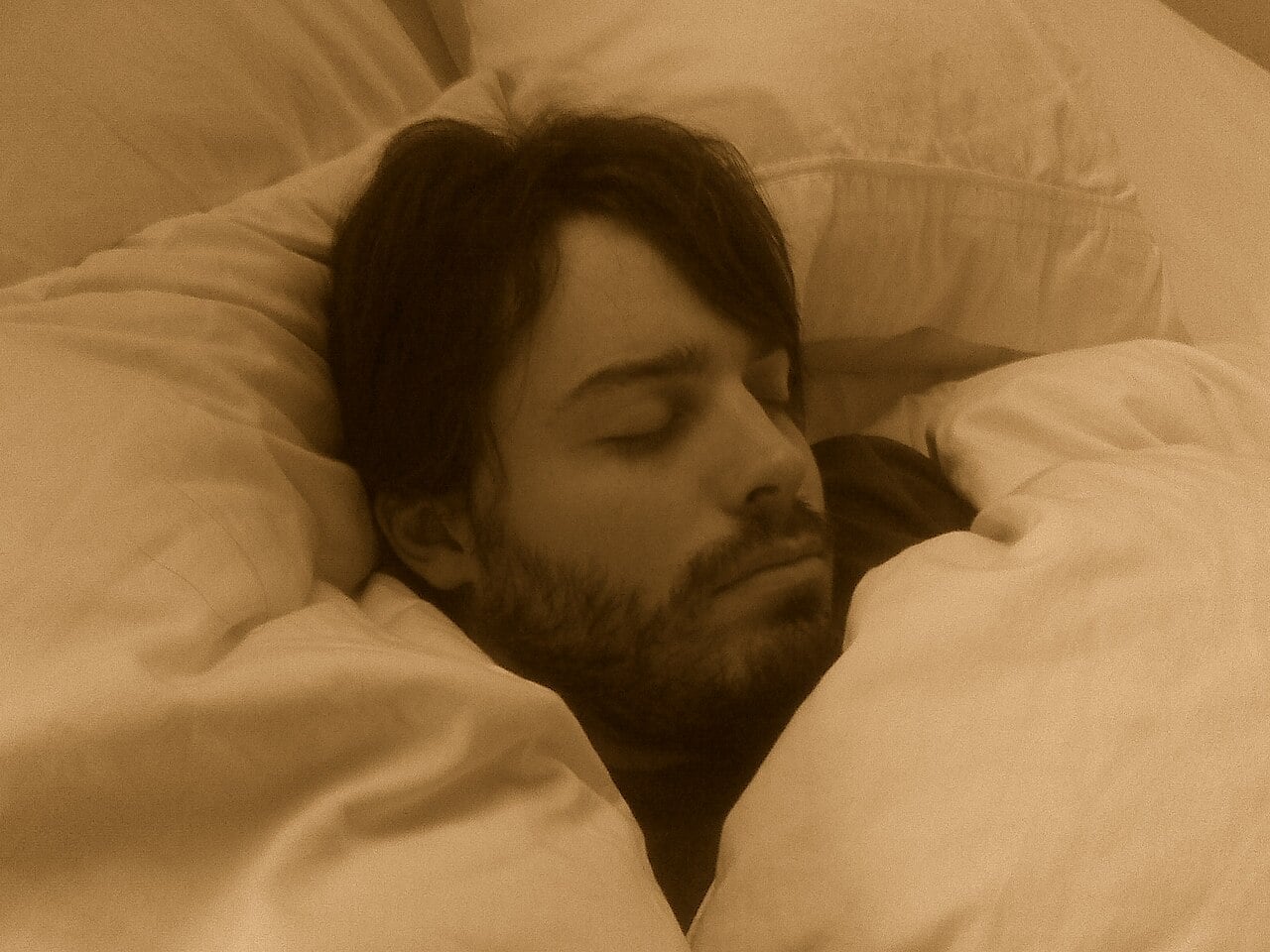 A bearded person with dark hair is sleeping peacefully in bed, surrounded by light-colored pillows and blankets. The image has a sepia tone.